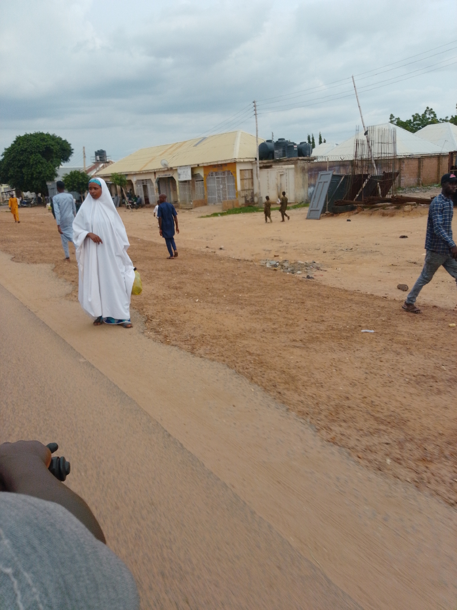 The image shows a street scene in Pantame, Nigeria, with several people walking along a dirt road. A woman in a white hijab and abaya walks towards the camera, holding a yellow bag. To her left, a man in a blue outfit walks away from the camera. Further in the background, other individuals are visible, along with buildings, trees, and construction materials. The sky is overcast with clouds. The foreground shows a portion of what appears to be a vehicle or person on a motorcycle, with a hand visible on the handlebars.