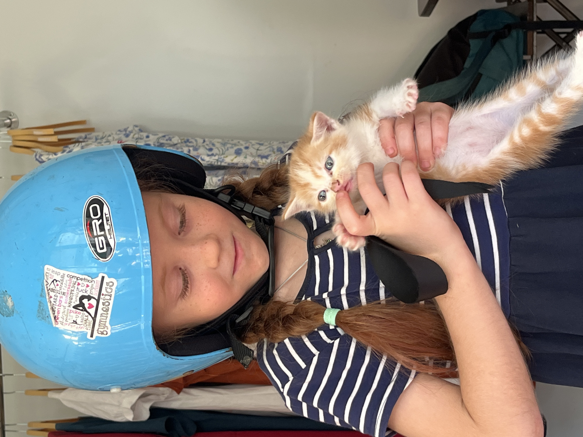 A young girl wearing a blue helmet and a navy and white striped shirt is holding an orange and white kitten. The girl has red hair in braids and freckles on her face. She is looking down at the kitten with a gentle expression. The kitten appears to be a few weeks old and has blue eyes. The background of the image shows a rack of clothes on hangers, suggesting an indoor setting. There is no indication of the location being Nha Trang, Vietnam.