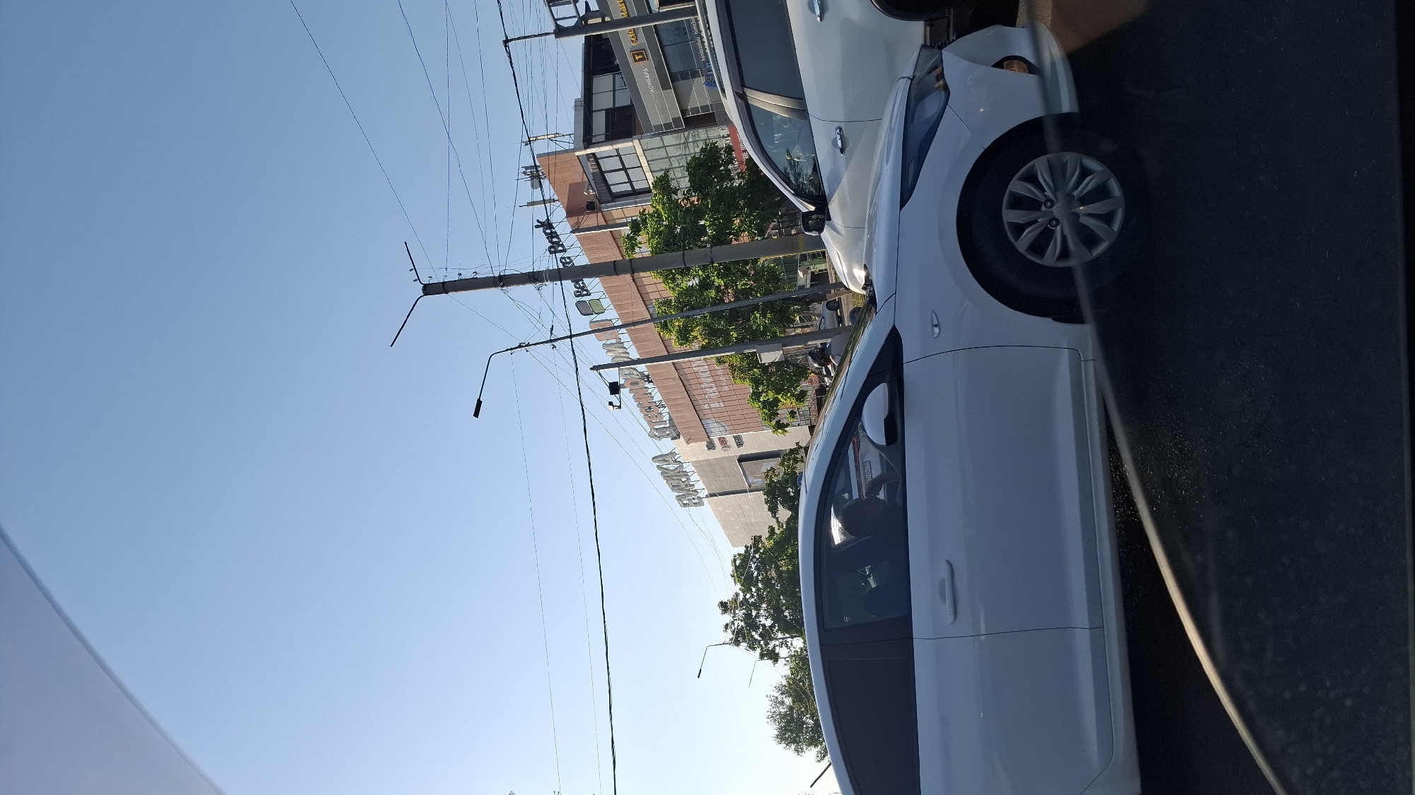 The image depicts a daytime scene on a city street, likely in Shymkent, Kazakhstan. A white sedan is in the foreground, partially obscured by the camera's perspective. In the background, a large, multi-story building with signage for "BEVERLY BANK" and "EVRIKA TECHNODOM" is visible. Trees line the street in front of the building, and a second white vehicle is parked or driving nearby. Overhead, power lines and streetlights are present. The sky is clear and blue.
