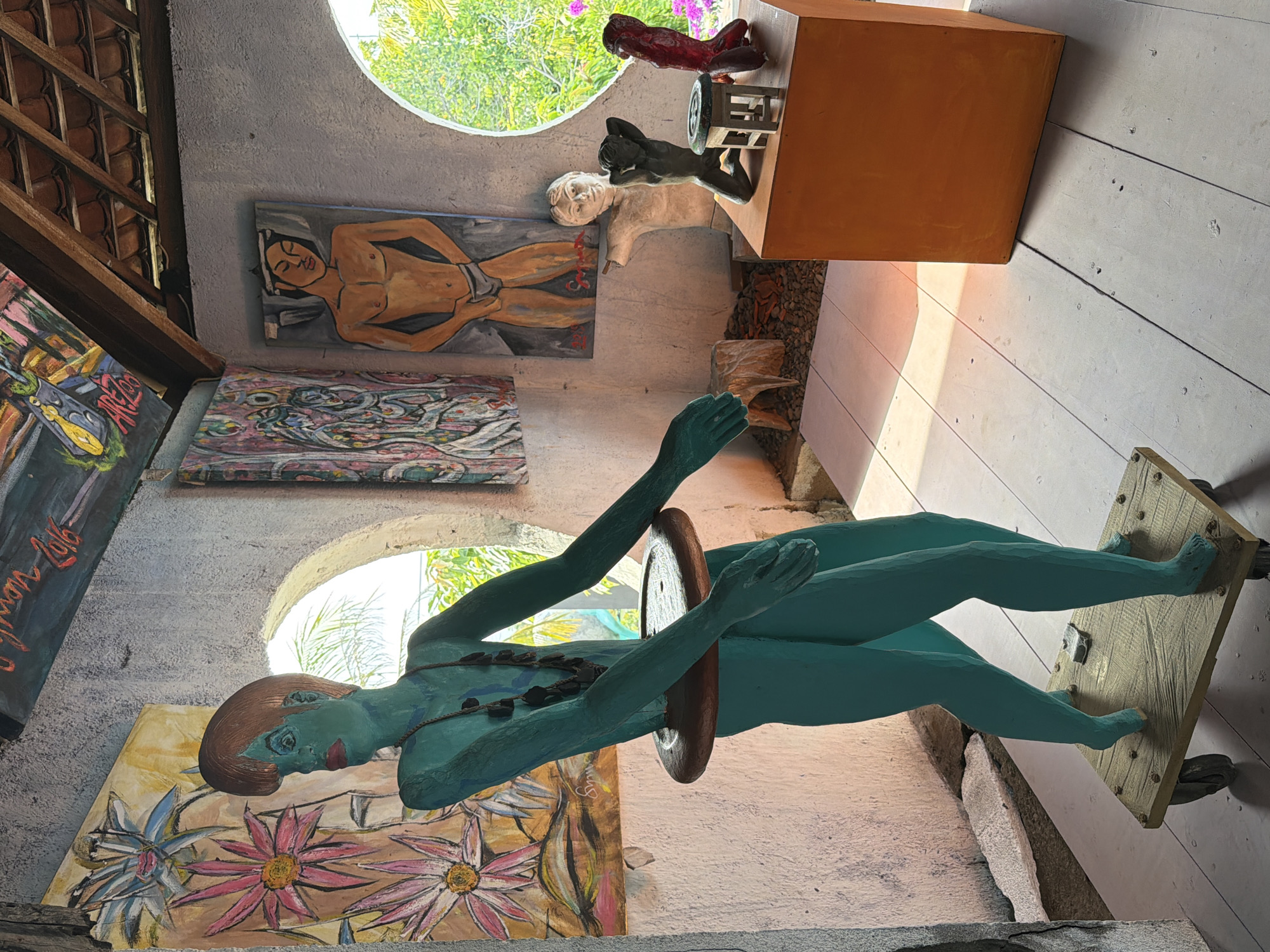 In Tejakula, Indonesia, within an artist's studio, a life-sized turquoise sculpture of a person stands center. The sculpture is mounted on a wooden platform with wheels and features a brown ring around its waist. It wears a dark beaded necklace. The studio walls are textured and painted, adorned with various artworks. To the left, a painting depicts stylized pink and yellow flowers. To the right, a circular opening reveals lush green foliage and trees outside. On a shelf behind the turquoise sculpture, a painting of a bare-chested man is displayed. In the background, on an orange pedestal, are three smaller sculptures: a pale bust of a person, a dark human figure in a kneeling pose, and a red abstract form. The floor is made of light-colored wooden planks. The ceiling, made of wooden beams and terra cotta tiles, is visible at the top of the image, with more artwork painted on the ceiling. The environment is filled with natural light, casting soft shadows.