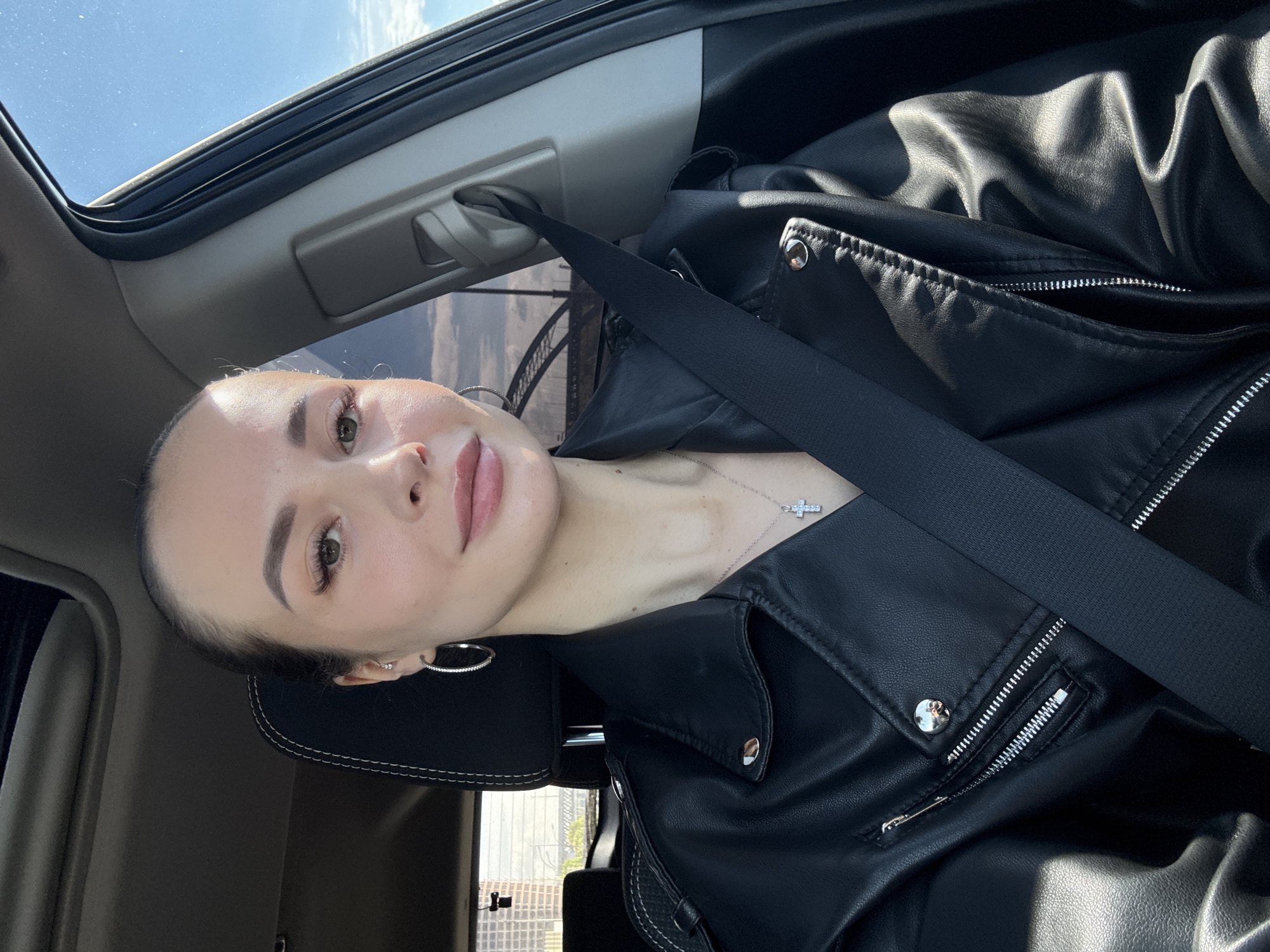 A woman is seated in a car, presumably in Kyiv, Ukraine, judging by the architecture visible through the open sunroof. She is wearing a black leather jacket and a seatbelt. She has her hair pulled back, is wearing hoop earrings, and a cross necklace. The interior of the car is light-colored. The sky is visible through the sunroof.