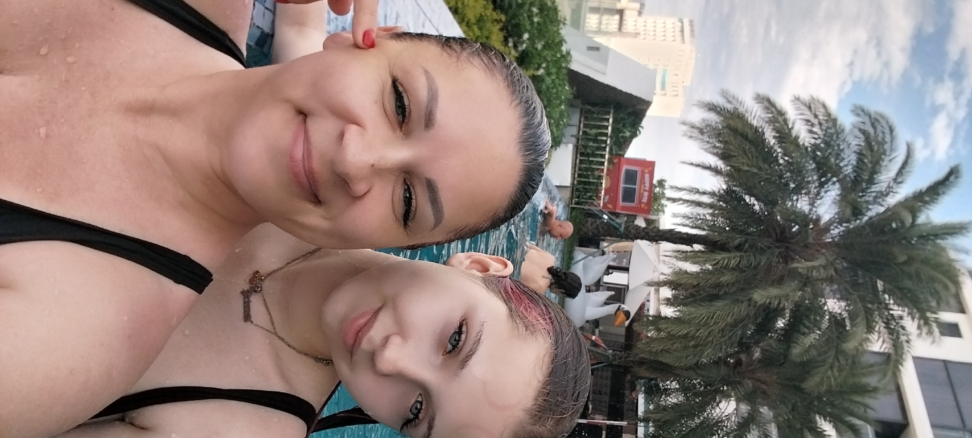 Two women are taking a selfie in a swimming pool, with a palm tree and buildings in the background. The woman in the foreground on the right has light skin, blue-green eyes, and light brown hair with pink streaks. She is wearing a black bikini. The woman behind her on the left has light skin, dark hair, and is wearing a black bikini top. She has red nail polish on her thumb.
The scene appears to be outdoors during the day. The sky is cloudy. In the background, there is a tall, modern building and a palm tree. There is also a small red structure with "HAPPY BITE" written on it. Other people are visible in the pool, as well as lounge chairs and umbrellas, suggesting a resort or hotel setting. The water is clear and blue. The overall mood appears relaxed and happy.