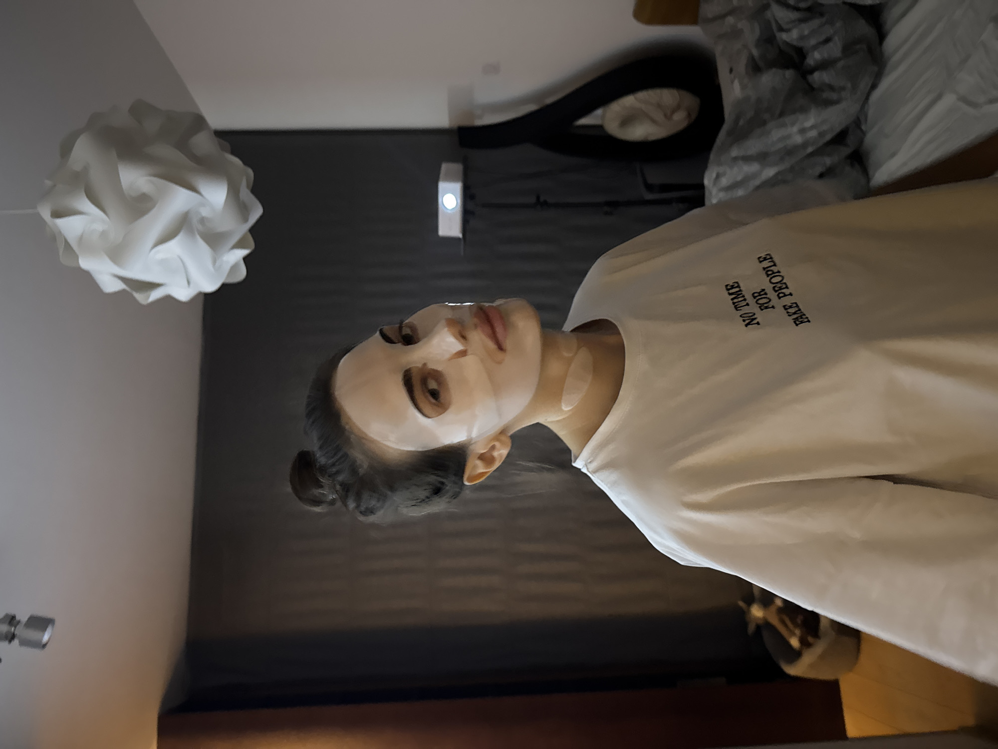 A woman is wearing a white T-shirt with the phrase "NO TIME FOR FAKE PEOPLE" printed on it. She is also wearing a facial mask. The room has a white decorative light fixture hanging from the ceiling and a gray wall. There are other objects in the background, including a dark-colored item hanging on the wall and items on a bed. The location is Unknown, Unknown.