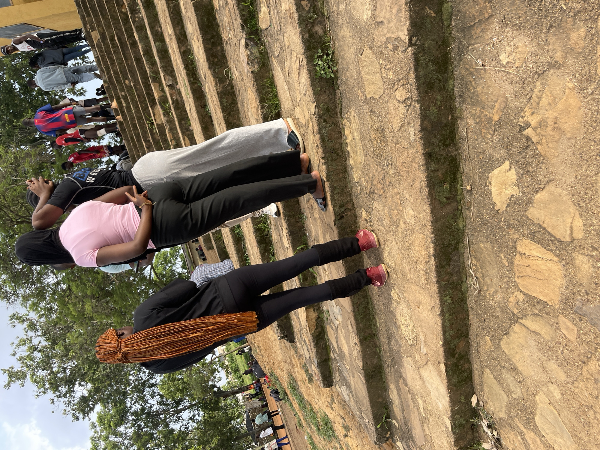 In Rafin Par, Nigeria, a group of people are gathered on a series of outdoor stone steps. In the foreground, a person with long, braided auburn hair is wearing a black top and black leggings, and red sneakers. To their right, another person in a pink shirt and black pants stands with their back to the camera. Further up the stairs, more individuals are visible, some standing, others sitting. The environment appears to be a park or public space, with trees and foliage surrounding the steps. The ground is a mix of dirt and stone. The overall scene captures a moment of casual gathering and interaction among the individuals.