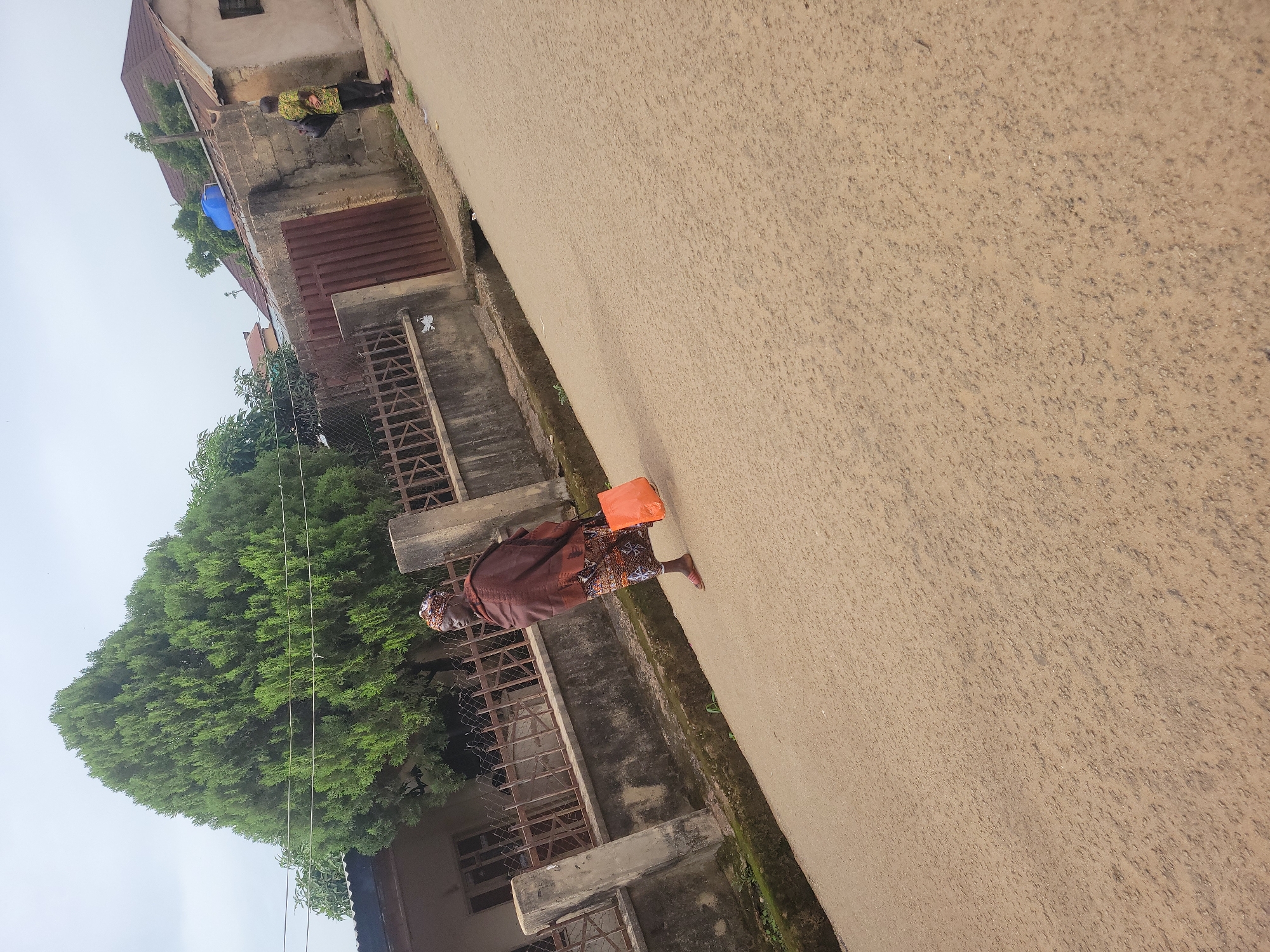 A medium shot captures an elderly woman walking along a paved street in Jos, Nigeria. She is dressed in a brown shawl and a patterned skirt, carrying an orange bag in her right hand. The street is light brown and appears somewhat uneven, with a gutter running along the left side. Behind her, a concrete wall topped with a metal fence lines the street. A large, dense green tree stands prominently behind the fence, casting shadows. Further in the background, a building with a brown roof and brick walls is visible, with another person standing near its entrance. The sky is overcast, suggesting a cloudy day.