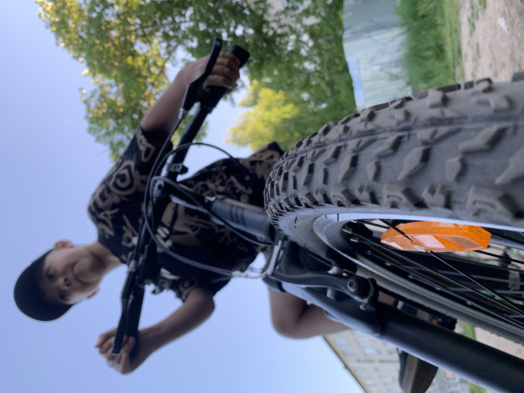 A low-angle, upward-facing shot features a young person riding a bicycle. The focus is on the front tire and the cyclist. The tire is black with a visible tread pattern and an orange reflector attached to the spokes. The cyclist, wearing a patterned black shirt and a black baseball cap, is holding the handlebars. The background shows a blue sky, trees, and a building suggesting an urban or suburban environment, possibly located in the city of Chaikovsky, Russia.