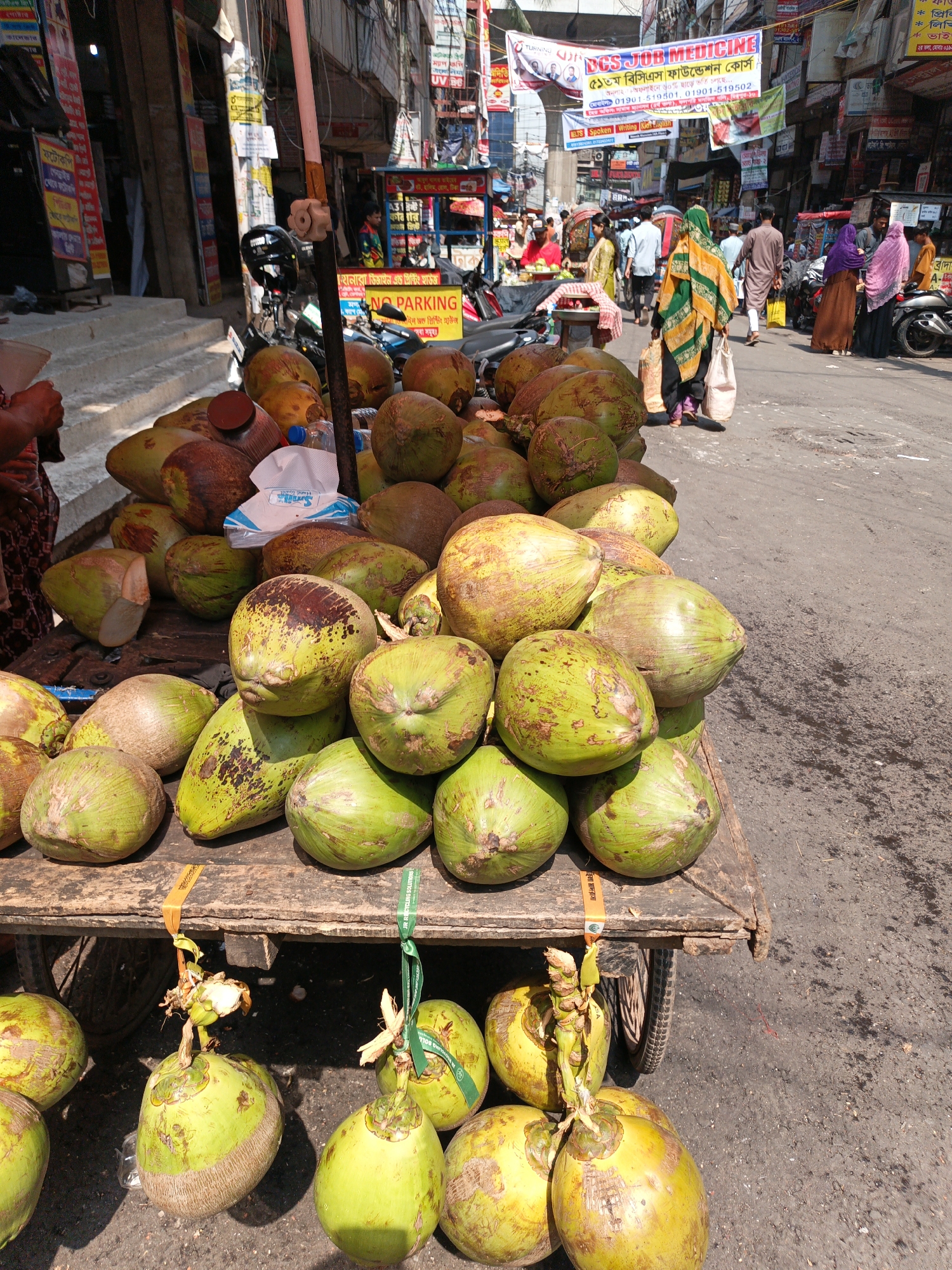 The image captures a vibrant street scene in Dhaka, Bangladesh. A wooden cart piled high with green coconuts is in the foreground. The coconuts vary in color, ranging from green to yellow. Some are decorated with ribbons and tags. Behind the cart, a crowded street bustling with people is visible. Many pedestrians are walking in both directions, and several are wearing traditional clothing, including headscarves and robes. Motorbikes and scooters are parked on the side of the road. In the background, numerous buildings with colorful advertisements and signs cover the façades. Banners are strung across the street overhead. Overhead electric wires crisscross above the street. The pavement is cracked and shows signs of wear. The image conveys a sense of the busy, urban environment in Dhaka.