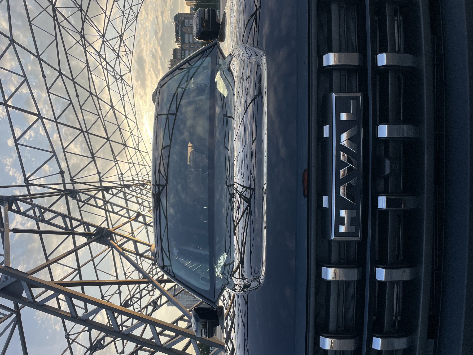 A black Haval SUV is parked in front of a large metal truss structure, possibly part of a bridge or stadium, with a cloudy sky in the background. The setting appears to be an urban environment, given the buildings visible in the distance. The low angle of the sun suggests it could be early morning or late afternoon. The vehicle's grille, with the Haval logo, is prominently displayed. The overall impression is a modern vehicle against an industrial or architectural backdrop. The location is identified as Kazan, Russia, though this specific detail cannot be visually confirmed from the image alone.
