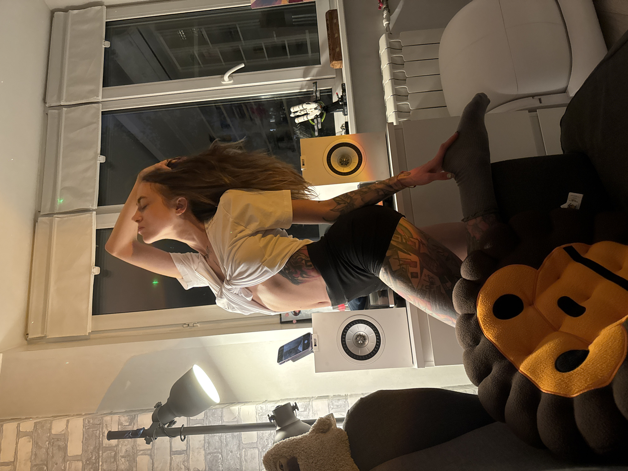 A woman with extensive tattoos on her legs and torso is kneeling on a chair in front of a window. She is wearing a white t-shirt tied at the waist, black shorts, and gray socks. Her left arm is raised behind her head, and her hair is down. Two white speakers are placed on a shelf behind her, one with a phone on top. The room is dimly lit, with a floor lamp to the left and a radiator on the wall. The window shows a dark exterior with some lights visible. A plush monkey-themed pillow is in the foreground, partially obscuring the woman's legs. The setting appears to be an indoor residential space.