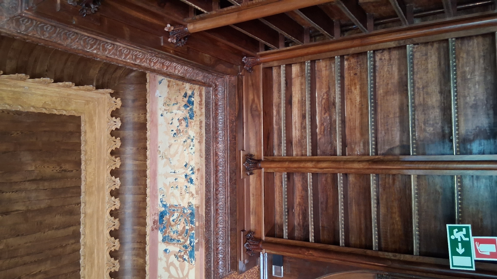 The image shows an interior view of a room, possibly a library or a study, characterized by extensive use of dark wood and ornate architectural details. The ceiling features exposed wooden beams and a decorative painted frieze. Below the ceiling, there are tall, wooden bookshelves or built-in cabinets with multiple shelves. The shelves are made of wood and appear to be empty. Two substantial wooden pillars with decorative capitals stand in the foreground, supporting the structure above. A green and white exit sign with an arrow pointing left and a running figure is visible on the lower left side of the image, next to a red fire extinguisher sign. The overall impression is one of historical grandeur and craftsmanship. This scene is likely located within one of the historic palaces or estates in Sintra, Portugal, given the architectural style and materials.