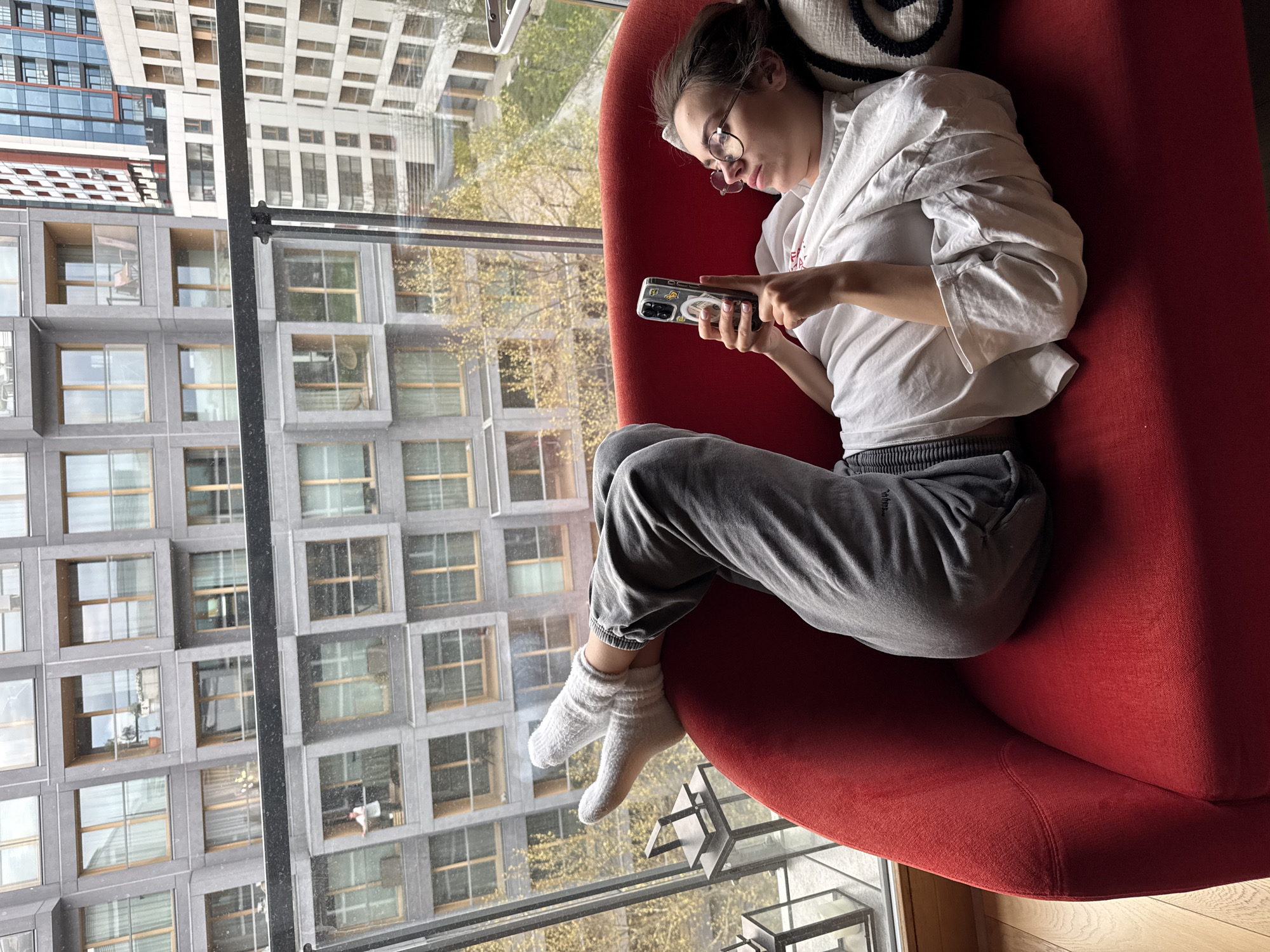 A young person reclines comfortably on a vibrant red armchair next to a large window, engrossed in their smartphone. The individual, appearing to be female, has dark hair pulled back, wears glasses, a white long-sleeved t-shirt with a faint red graphic, gray sweatpants, and white socks. They are holding a smartphone with a clear case adorned with several stickers, scrolling or typing with both hands. Behind their head, a white cushion with a black spiral pattern is visible.
The scene is set indoors, looking out onto a modern urban landscape. Through the window, several multi-story buildings are visible under what appears to be clear daylight. One prominent building has a distinctive grey facade with numerous square, recessed windows. Another building, further away, features lighter-colored walls and more traditional rectangular windows, with a section of blue-tinted reflective glass also visible. Patches of green and yellow foliage from trees suggest a spring or autumn setting. The overall perspective is slightly from above, looking down into a city street or courtyard between buildings, consistent with a view from a higher floor in an apartment or office building in Warsaw, Poland. The person seems relaxed and absorbed in their digital activity. No explicit text is clearly legible on the clothing or phone stickers, beyond indistinct patterns.
