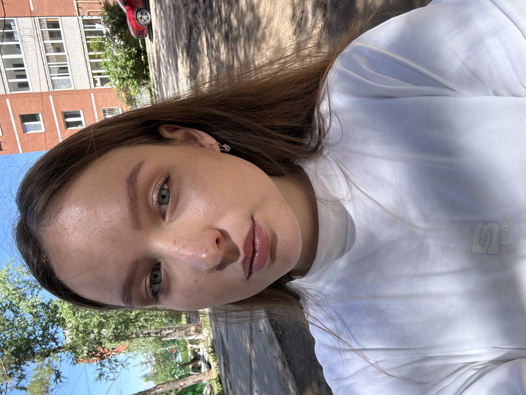In an outdoor urban environment, a young Caucasian female with long brown hair, fair skin with freckles, and blue eyes is photographed from the chest up. She is looking directly at the camera and wearing a white t-shirt with a subtle logo on the left chest. She has a small earring in her left ear. The background includes a brick apartment building, trees with green foliage, and a paved area with shadows cast from overhead trees. The lighting suggests it is a sunny day. The building in the background is identifiable as an apartment building located in the city of Chaikovsky, Russia.