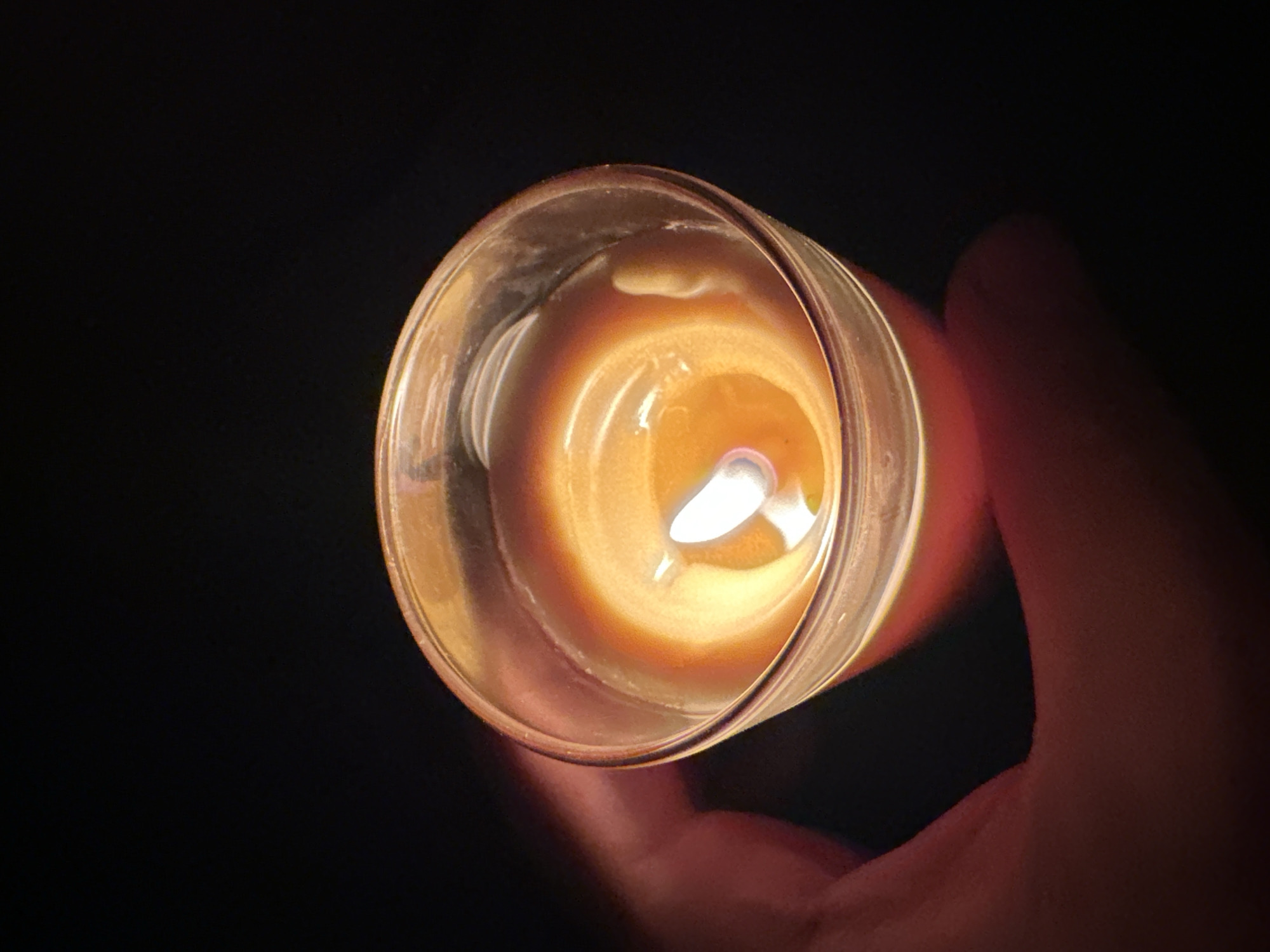 A lit candle in a glass jar is held by a hand against a dark background. The candle flame is bright white with an orange glow. The melted wax of the candle forms a wavy pattern. The hand is out of focus, with warm skin tones visible. The scene is dark, suggesting it is nighttime or indoors with minimal lighting. The location in Comrat, Moldova is not identifiable from the image itself.