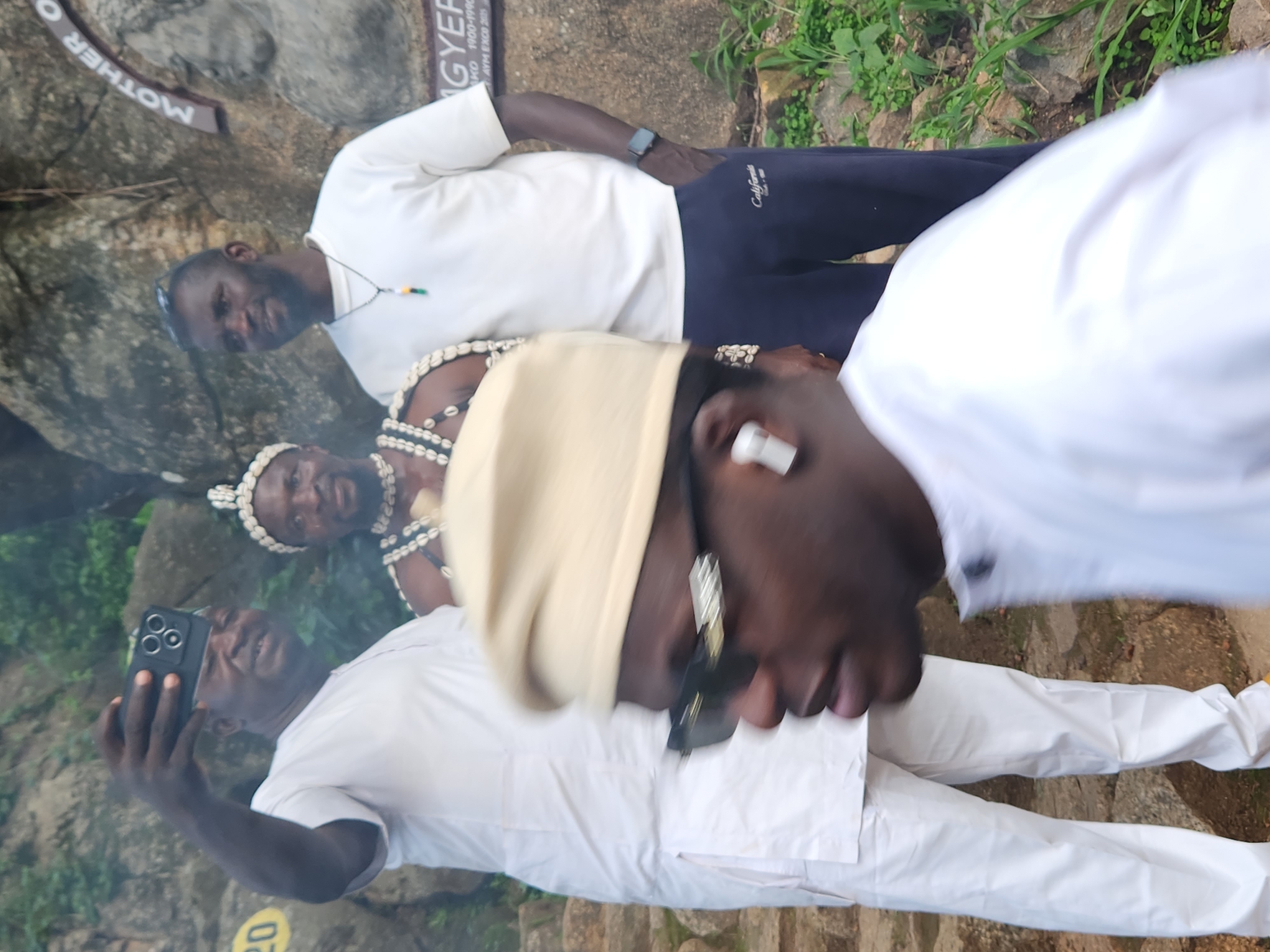 The image shows a group of men, likely in Jos, Nigeria. One man in the foreground, wearing a white outfit and a tan head covering, is taking a selfie with a smartphone. He is blurred due to motion. Behind him, two other men are visible. One is wearing a white long-sleeved shirt and dark pants, and appears to be looking at the camera. The second man behind him is wearing traditional attire with necklaces and a headpiece, and is also looking towards the camera. In the background, there are rocks and greenery, suggesting an outdoor setting. A sign with text is partially visible on the rocks to the left, with the words "MOTHER O" and "GYER" legible. There are also some yellow markings on the ground that are not fully decipherable. The lighting suggests it is daytime. The men appear to be in a casual gathering, possibly a tourist attraction or a cultural site.