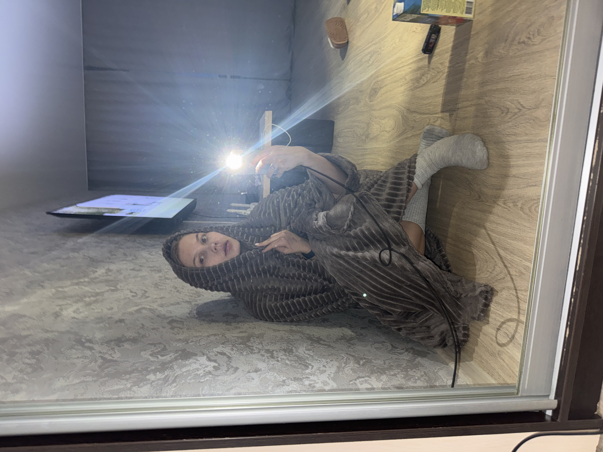 A person wrapped in a striped, gray blanket is lying on a gray and white patterned surface resembling a bed or the floor. They are holding a small, bright light source, possibly a phone flashlight, directed towards the camera. The person is wearing gray socks and has a serious expression. In the background, there is a television on a stand, and a wood-grain floor with miscellaneous objects including a shoe, a box, and a small black remote control. A window frame is visible around the edge of the image. The exact location within the city of Chaikovsky, Russia, cannot be determined from the given information.