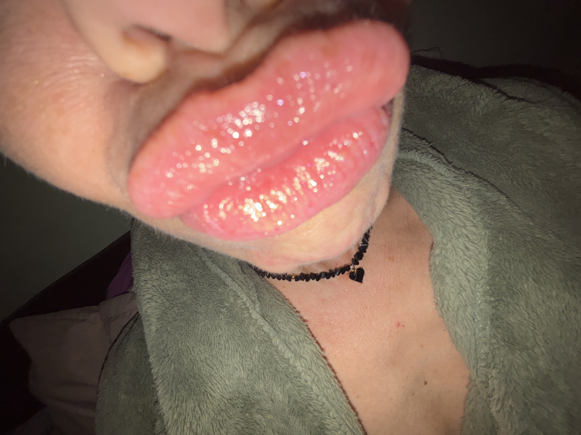 The image is a close-up of a person's face, focusing on their lips and chin. The lips are plump and appear to have lip gloss on them, reflecting light. The person is wearing a fuzzy, olive-green robe or blanket. A black beaded necklace with a small black heart pendant is visible around their neck. The background is dark and out of focus, suggesting an indoor, dimly lit environment. There is no indication of location in the city of Unknown, Russia.
