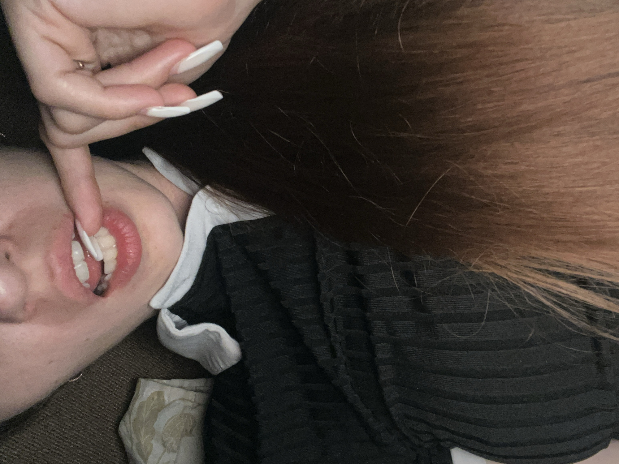 A close-up, over-the-shoulder shot shows a person lying down, with their head tilted to the side and their hair spread out. Their right hand, with long white acrylic nails, is near their face, with a finger touching their lips. The person is wearing a black ribbed top with a white collar. The background is a brown textured surface, possibly a couch. The lighting is dim, with a soft glow on the skin and hair. The image appears to be taken indoors, in a domestic setting, and there is no indication of the city or specific location.