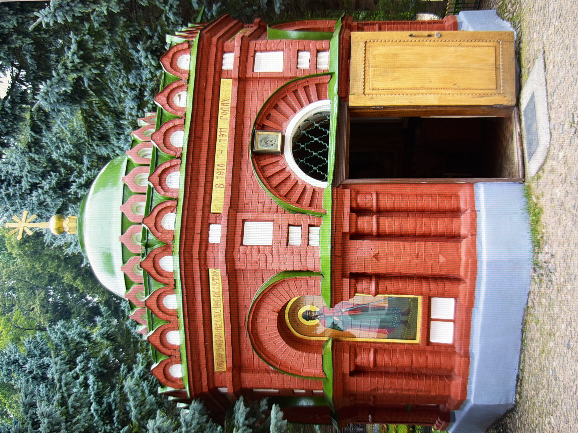 This is a medium-shot, eye-level photograph of the Church of the Savior at the Kyiv Pechersk Lavra, located in Kyiv, Ukraine. The church is a red brick building with a green roof and a golden dome topped with a cross. The church has multiple arched windows and decorative elements. There is an icon of a saint displayed in an alcove on the left side of the building, and an open wooden door on the right side, revealing a dark interior. The church is surrounded by lush green trees. The ground in front of the church is paved with cobblestones. The overall scene depicts a religious building in a serene, natural setting.