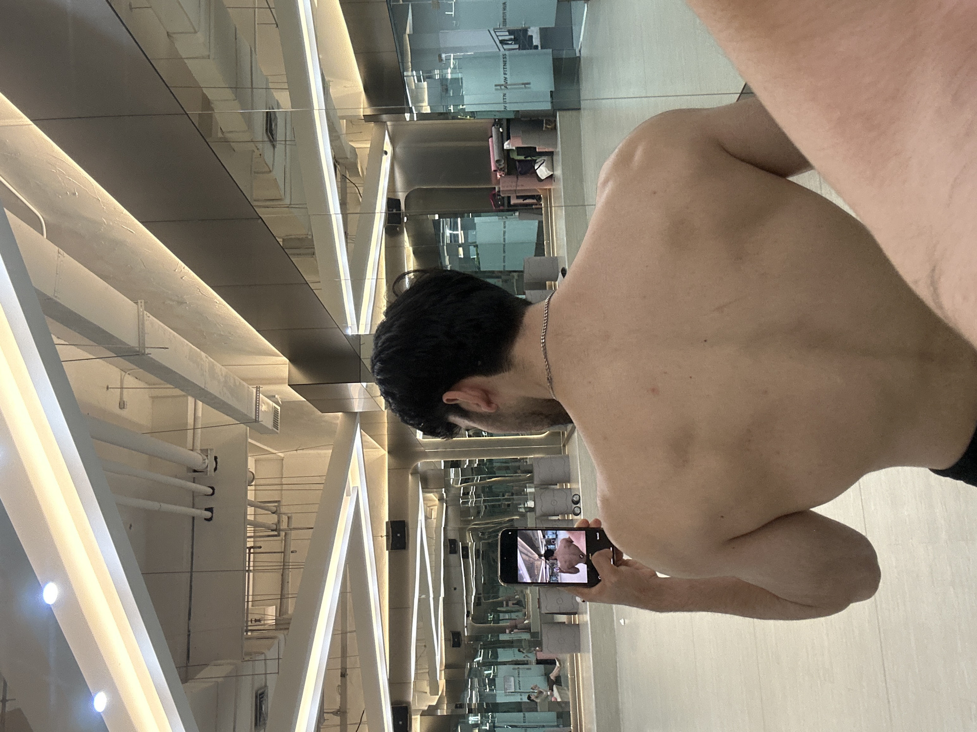 The image, rotated 90 degrees clockwise, depicts a shirtless man viewed from his back and right side, standing in a modern indoor environment characterized by extensive mirrored or highly reflective surfaces. He has dark hair and is wearing a silver chain around his neck. The man is holding a smartphone in both hands, positioned to capture an image or video, with his own reflection visible on the phone's screen.
The setting appears to be a contemporary building, possibly a fitness center or commercial space, featuring prominent white, angular structural elements that may be beams or pillars, equipped with integrated strip lighting. These reflective surfaces create multiple reflections of the ceiling, which reveals exposed pipes, ventilation ducts, and other industrial fixtures.
Through the reflections on glass partitions in the background, the text "HVN FITNESS" is visible, confirming the location as a gym or fitness facility. Other reflections show glimpses of interior spaces with various objects, possibly gym equipment or yoga mats. The floor is a light-colored, tiled surface. The lighting throughout the space is bright and artificial. The scene takes place in the Chongqing High-tech Zone, China.