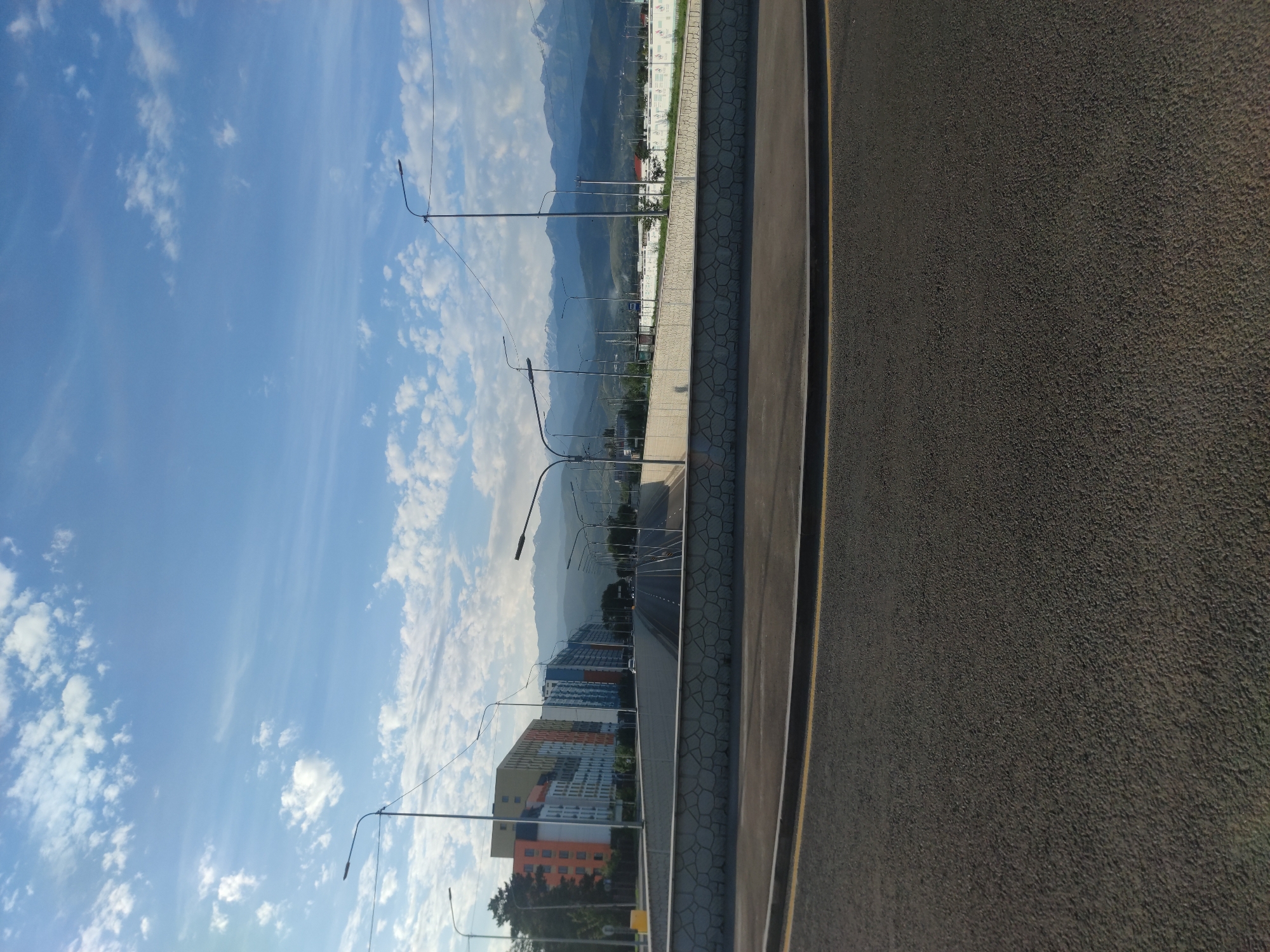 The image captures a view of a highway underpass in Almaty, Kazakhstan, with modern apartment buildings visible on the left and a majestic mountain range in the background. The sky is a clear blue with scattered white clouds, suggesting fair weather. The foreground shows a section of asphalt road with yellow lane markings. The underpass itself features a concrete retaining wall with a decorative stone pattern. Streetlights line the roadway leading into the underpass. The apartment buildings are contemporary in design, featuring a mix of colorful facades. The mountains in the distance are snow-capped, a characteristic feature of the Almaty landscape. There are no people visible in the image, and the scene appears to be a quiet, urban environment with a natural backdrop.