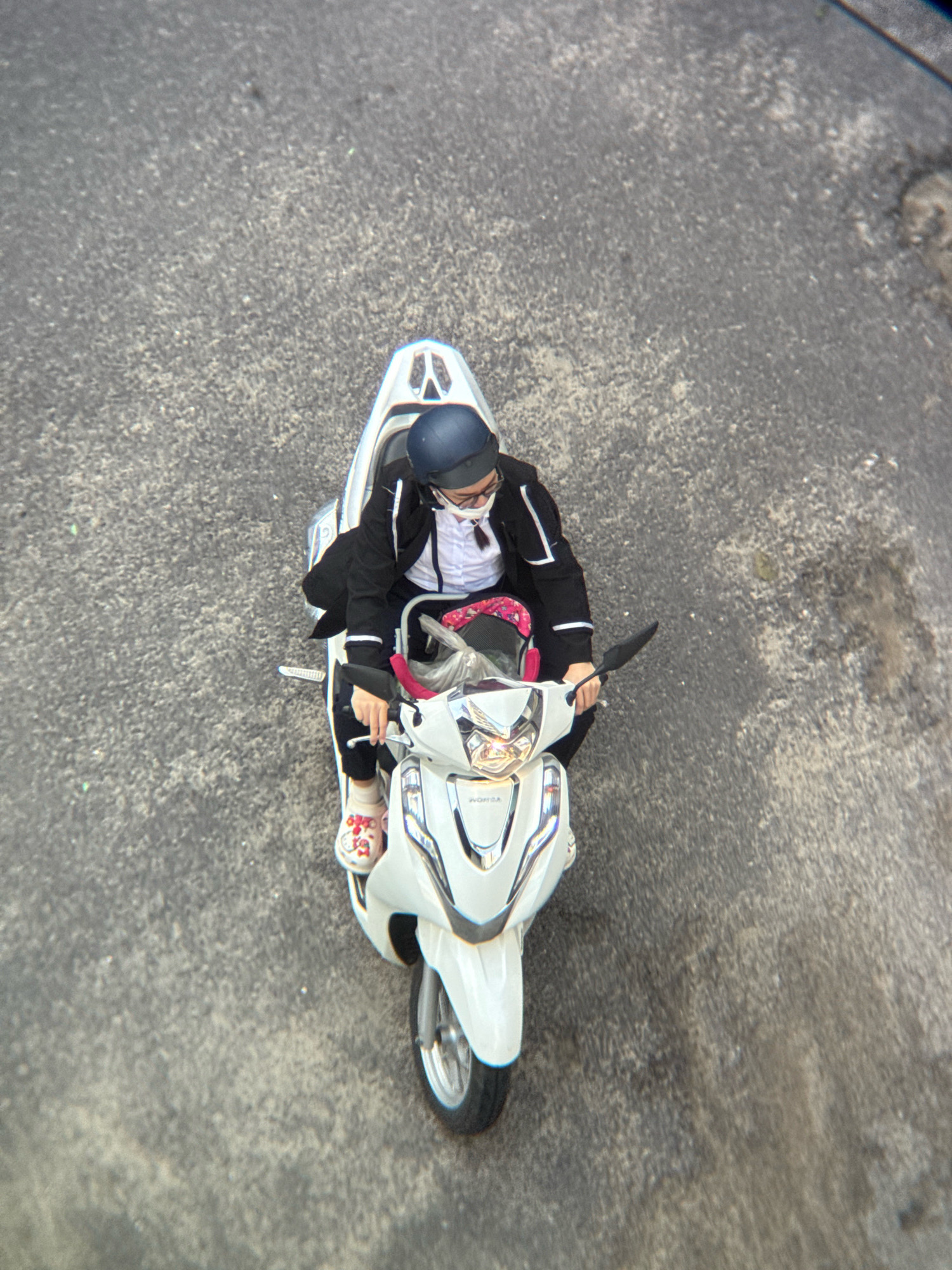 A person wearing a dark blue helmet, white face mask, glasses, a white shirt, a black jacket with white trim, and dark pants rides a white Honda scooter on a grey asphalt road. The person is wearing light-colored clogs with a cartoon character design. The scooter has its headlights on. The scene is an aerial view, likely taken from an elevated position. The road surface is textured and appears slightly worn. The image context suggests an urban environment, potentially a street in Nha Trang, Vietnam, given the prevalence of scooter usage in such locations.