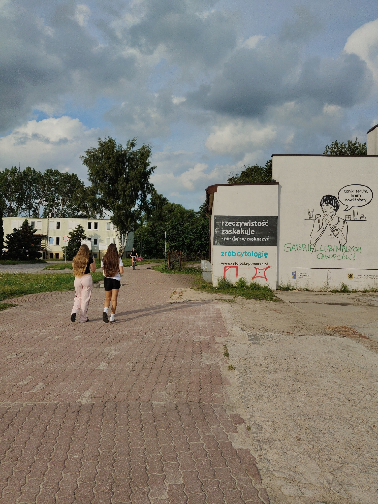 Two young women with long hair walk away from the camera on a red brick paved walkway. The woman on the left wears pink sweatpants and a black top. The woman on the right wears black shorts and a white t-shirt. Both are wearing black shoes and white socks. In the background, a person rides a bicycle. A white building with many windows is visible behind them, surrounded by trees. To the right of the walkway, there is a white building with graffiti. The graffiti includes a drawing of a woman with bottles of skincare products, text that reads "tonik, serum, krem nawilżający", and the words "GABRIEL LUBI MALYCH CHLOPOW!". Below this, there are logos for "Fundacja Pomorskie" and "URZAD MARSZAŁKOWSKI WOJEWODZTWA POMORSKIEGO". Another section of the building has a black sign with white text that reads "rzeczywistość zaskakuje -nie daj się zaskoczyć" and "zrób cytologię www.cytologia-pomorze.pl". The sky above is cloudy with patches of blue sky. The image appears to be taken in Rumia, Poland, given the visible signage.