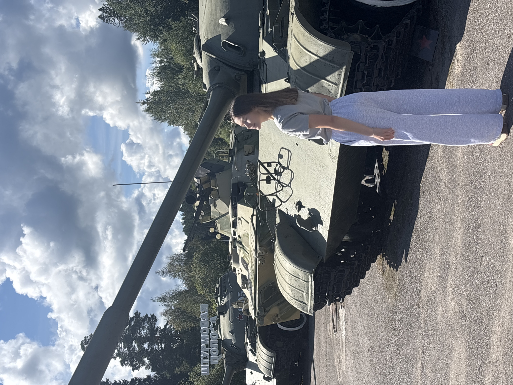 A young woman stands to the right of the frame, facing left and looking up at the sky. She is wearing a grey t-shirt and light grey sweatpants. Behind her and to her left are several military tanks, some of which are partially obscured. The tanks appear to be parked outdoors in what looks like a museum or historical site. Above the tanks, a sign with white letters is visible, reading "ШТУРМОВАЯ ПОЛОСА," which translates to "Assault Course." The environment is outdoors with trees and a cloudy sky. The location is identified as Gamay, Russia.