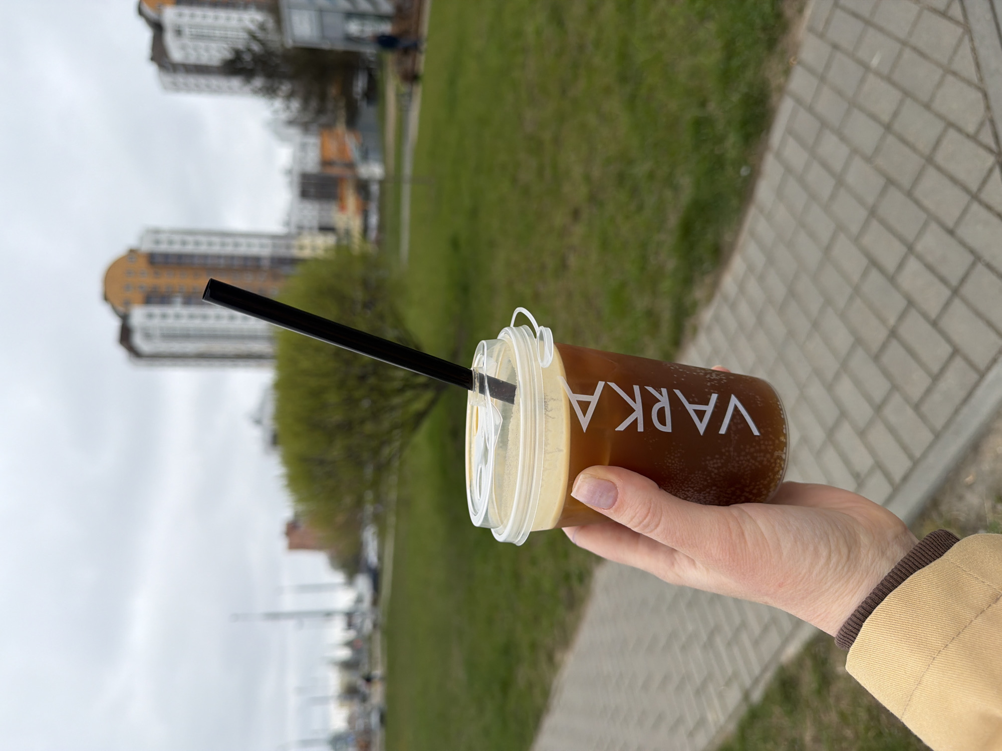 The image, set in Hrodna, Belarus, captures a first-person view of a hand holding a clear plastic cup filled with a dark brown beverage, likely coffee or tea, topped with a light-colored frothy layer. A black straw protrudes from the clear plastic lid. The word "VARKA" is visibly printed vertically in white capital letters on the side of the cup. The hand, with neatly kept nails, emerges from the sleeve of a beige or tan jacket, which has a contrasting dark brown ribbed cuff.
The scene depicts an outdoor urban environment, possibly a park or a pedestrian walkway alongside a street. To the right, a paved path made of light-colored rectangular blocks recedes into the distance. Adjacent to the path on the left, there's a wide expanse of green grass. In the blurred background, several multi-story apartment buildings with white, orange, and grey sections are visible, suggesting a residential area. Sparse trees, some showing early signs of foliage, are scattered across the green space. The sky is overcast, indicating a cloudy daytime. The activity is a person enjoying a takeaway drink while outdoors.