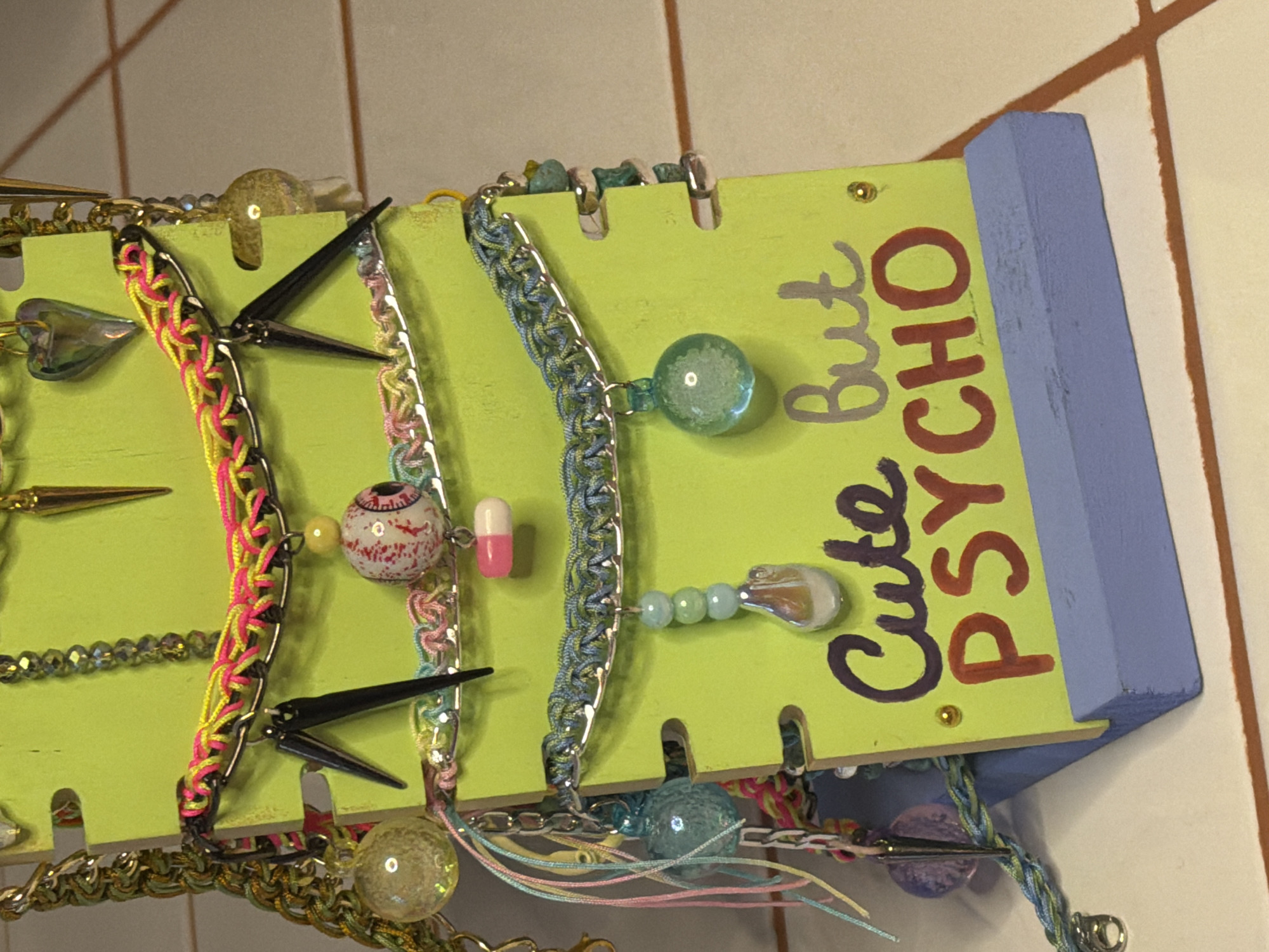 The image shows a lime green jewelry display stand with "Cute But PSYCHO" written in purple and red. The stand has a light blue top edge and is holding several handmade necklaces or bracelets. The jewelry includes chains, beaded strands, and charms, like a stylized eyeball and a pink pill. The background is a wall with beige tiles. The image does not contain information to identify the location in Belgrade, Serbia.