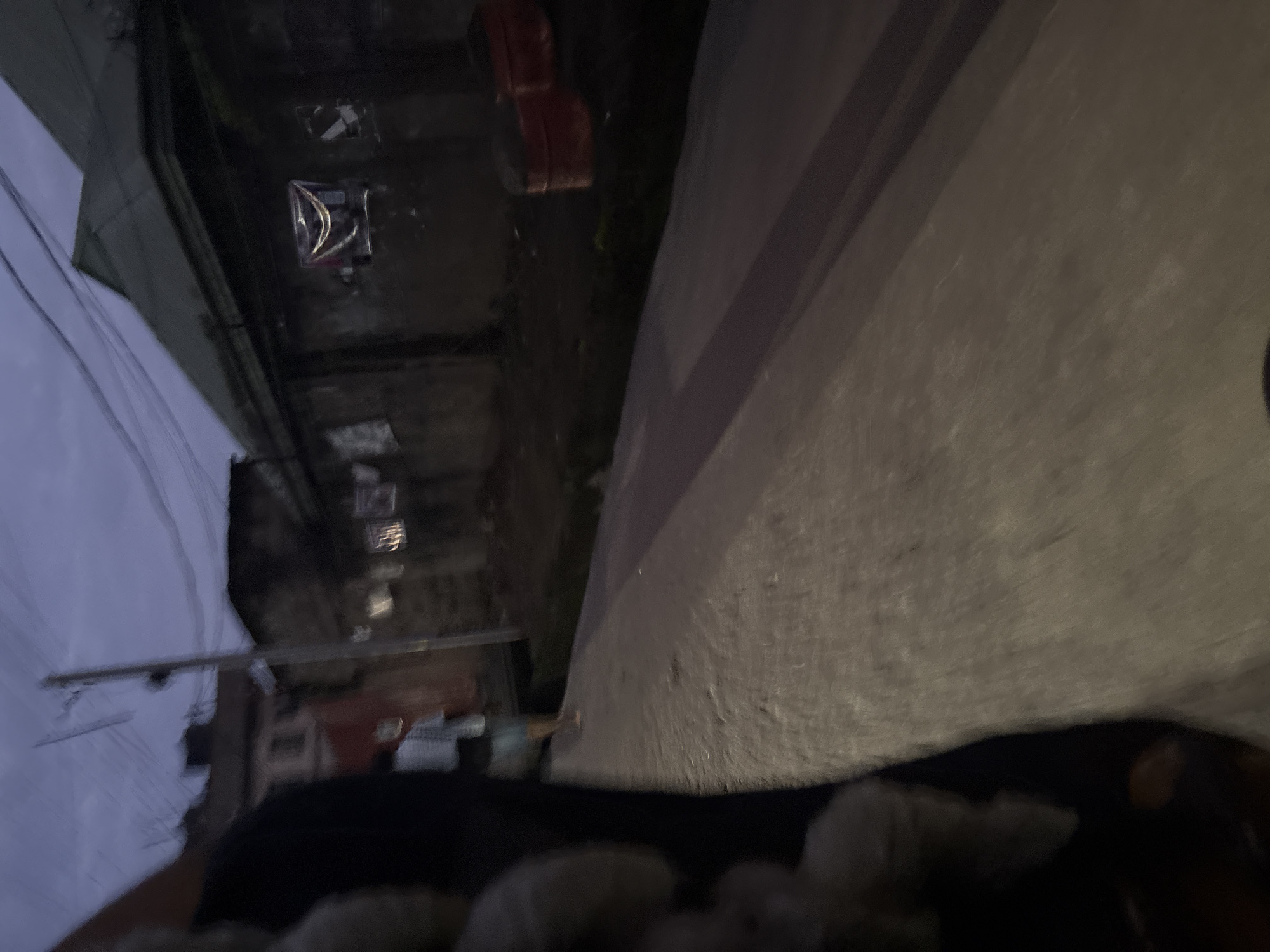 An overcast, dusk scene unfolds on a dusty street in Onitsha, Nigeria. The foreground is dominated by a blurred, dark object, likely a person's arm or leg, positioned to the left of the frame, creating a sense of motion. To the right, a person in a light-colored shirt and shorts walks away from the camera down a pathway that bisects the image diagonally. The pathway is a mix of compacted earth and scattered debris.
In the middle ground, a weathered wall runs horizontally across the image, adorned with posters or notices that are largely indistinguishable due to the low light and motion blur. Above the wall, the roofs of buildings with corrugated metal are visible. To the far right, two large, red, cylindrical objects, possibly drums or planters, sit on the ground next to the wall. A utility pole with a tangle of wires stands near the center of the frame. The overall impression is one of a residential area in the late evening, characterized by utilitarian structures and a somewhat unkempt environment.