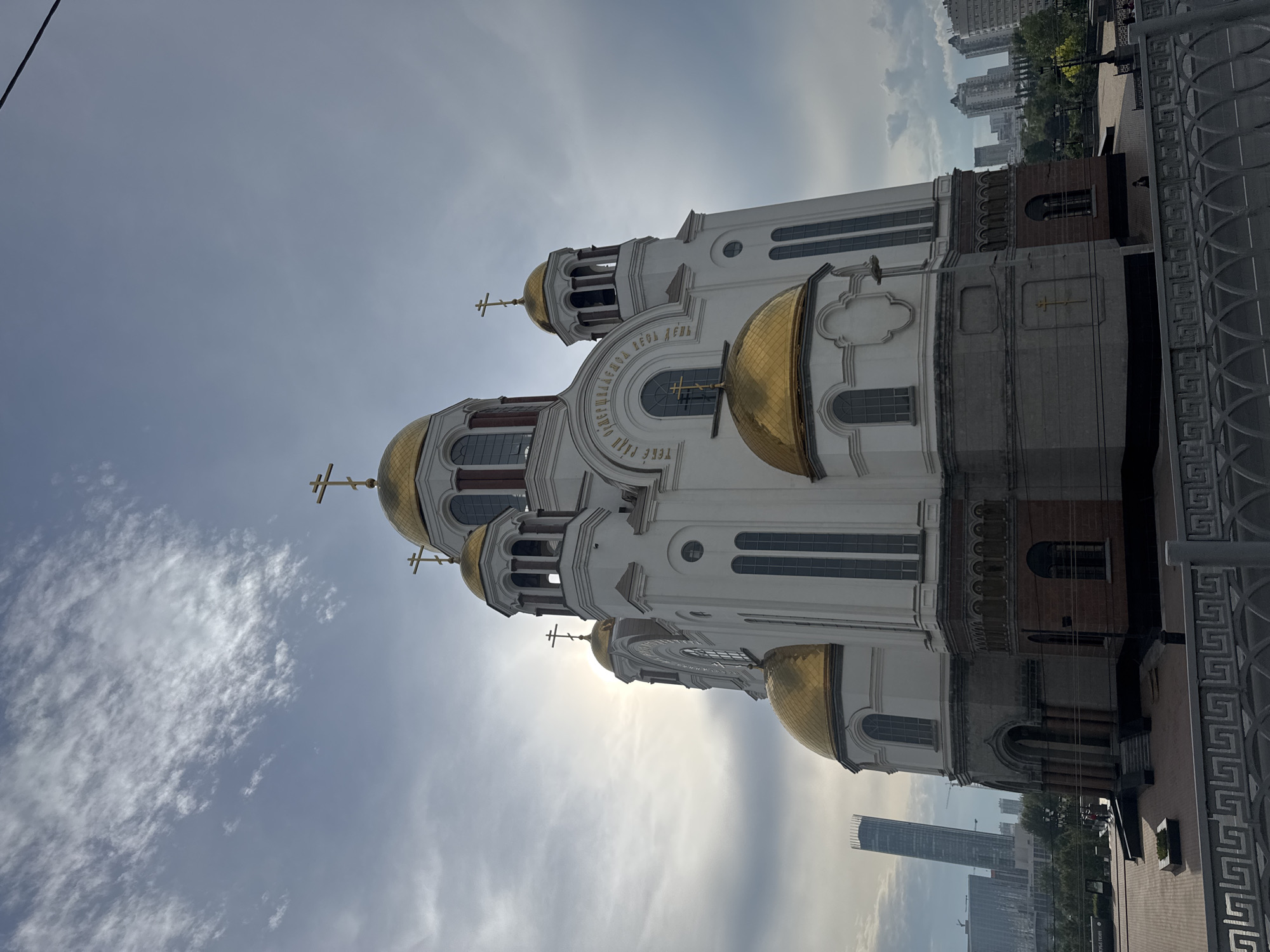 The image captures the Church on Blood in Yekaterinburg, Russia, under a cloudy sky. The sun is peeking through the clouds, creating a bright halo effect behind the central dome. The church, a prominent landmark, is white with golden domes topped with crosses. The architectural details, including arched windows and decorative elements, are visible. In the background, the modern cityscape of Yekaterinburg is visible, with tall buildings and skyscrapers contrasting with the historic church. The foreground shows a paved plaza and a decorative metal railing. The overall scene depicts a blend of religious architecture and urban development.