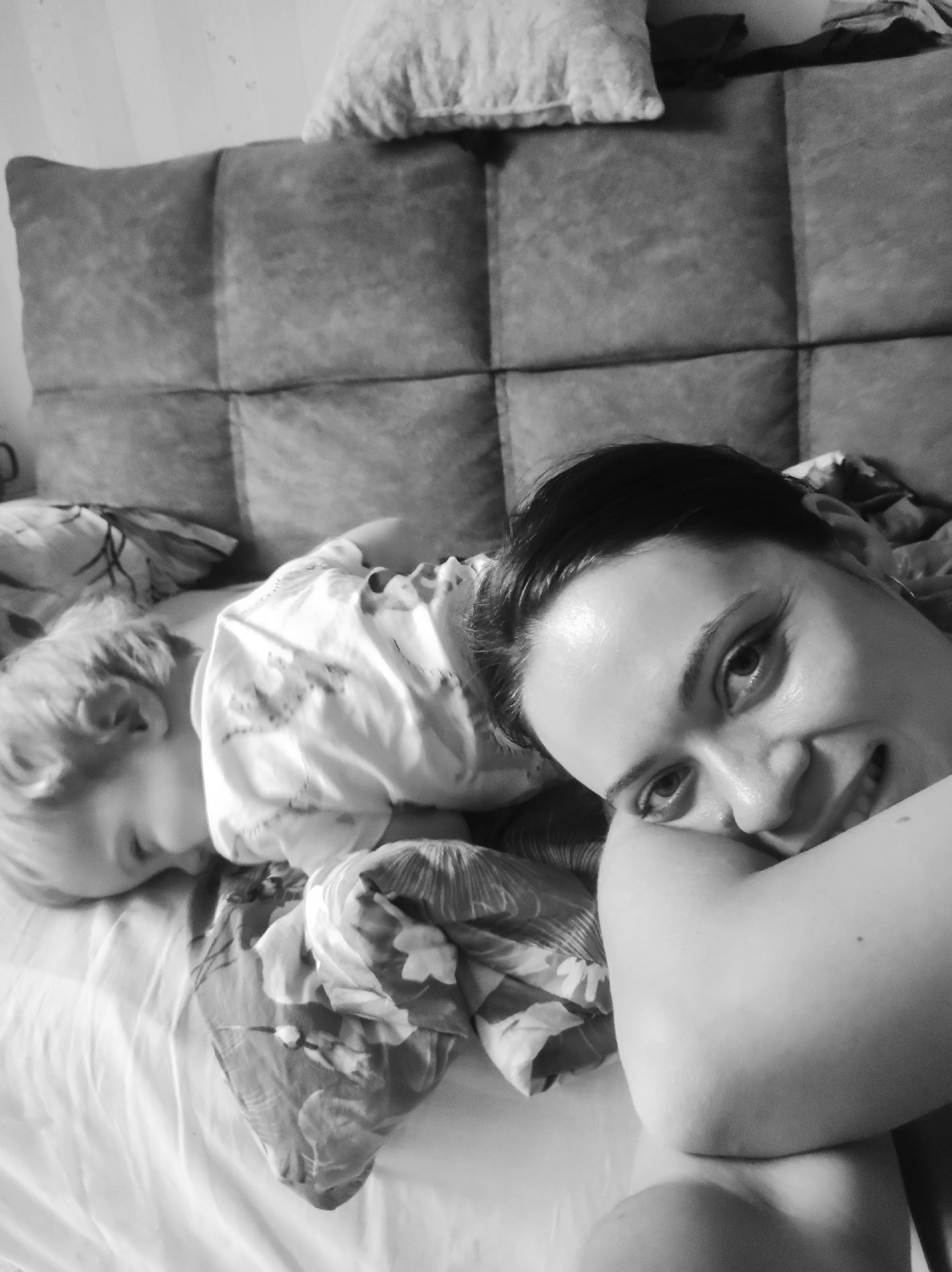 This black and white photograph, taken in Chaikovsky, Russia, captures an intimate indoor scene featuring a woman and a young child resting on a bed.
In the foreground and to the right, a woman is depicted from the chest up, smiling gently and looking directly into the camera. Her dark hair is styled neatly, and her facial expression conveys warmth and contentment. She appears to be lying on her side, with her left arm bent and her elbow visible, possibly propping herself up.
To her left, a young child, likely a toddler, is visible in the middle ground, appearing to be asleep or deeply resting. The child's light-colored hair, the back of their head, and a portion of their face and ear are discernible. They are wearing a light-colored, patterned top or sleepwear. The child is lying on a bed covered with white sheets and patterned bedding.
The background features a dark, padded, tufted headboard, indicating a bedroom setting. Above the headboard, a textured pillow and a subtly patterned wall are visible. The lighting is soft and even, contributing to a tranquil and peaceful atmosphere. The image depicts a quiet moment of familial closeness. There is no visible text.