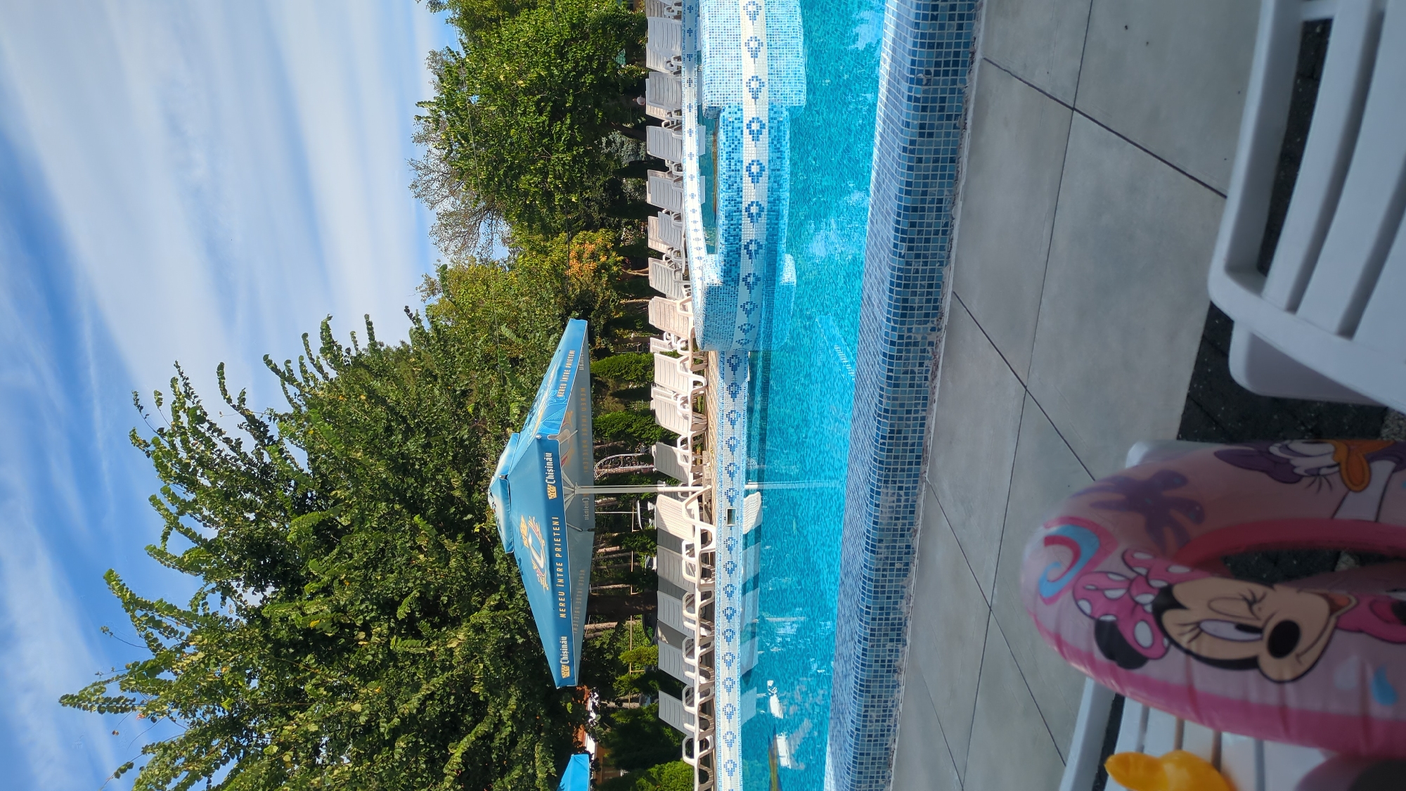 The image depicts an outdoor swimming pool area. A large, blue umbrella with the "Chisinau" logo and text is prominently displayed over a row of white lounge chairs. The pool itself is tiled with blue mosaic patterns. Lush green trees and vegetation surround the pool area, and a bright blue sky with wispy clouds is visible above. In the foreground, a pink inflatable ring featuring cartoon characters, including Minnie Mouse and Daisy Duck, rests on a white lounge chair. The scene appears to be set in the city of Vadul lui Voda, Moldova, suggesting a resort or recreational facility.