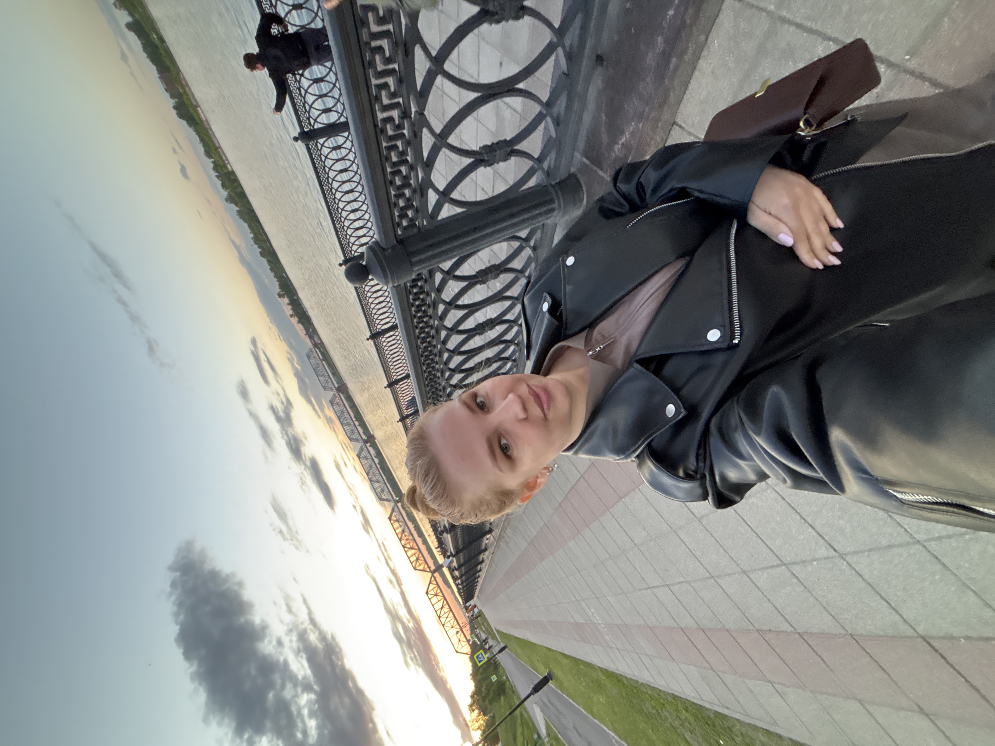 A young woman in a black leather jacket stands on a paved walkway next to a decorative wrought iron railing. In the background, the Nerskaya River reflects the colors of the sunset. A person is visible leaning over the railing in the distance. This location is likely in Rusanovo, Russia.