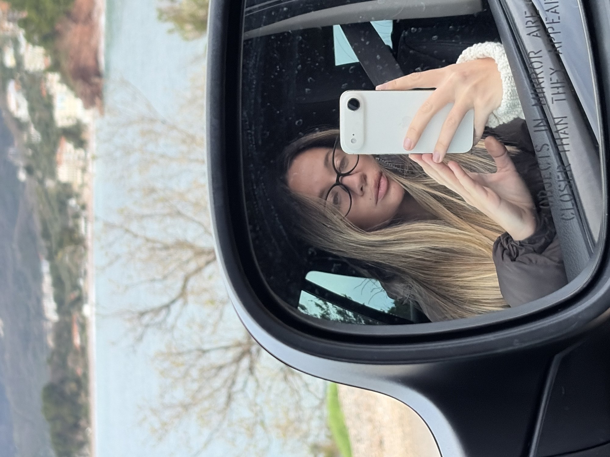 A photograph captures the reflection of a light-skinned female in a car's black-framed passenger-side mirror. The subject, with long blonde hair and black-framed glasses, wears a dark padded jacket and a light-colored knitted cuff, and is secured by a black seatbelt. She is actively taking a selfie using a white smartphone, which she holds with both hands, positioned to capture her reflection in the mirror. The mirror's lower right edge displays the partially visible warning "OBJECTS IN MIRROR ARE CLOSER THAN THEY APPEAR." Beyond the car mirror, the background depicts a coastal landscape in Unknown, Unknown, featuring a wide body of light blue water, a reddish-brown shoreline, and distant hills populated with numerous white buildings and red roofs. Bare tree branches are visible in the immediate foreground between the vehicle and the water.