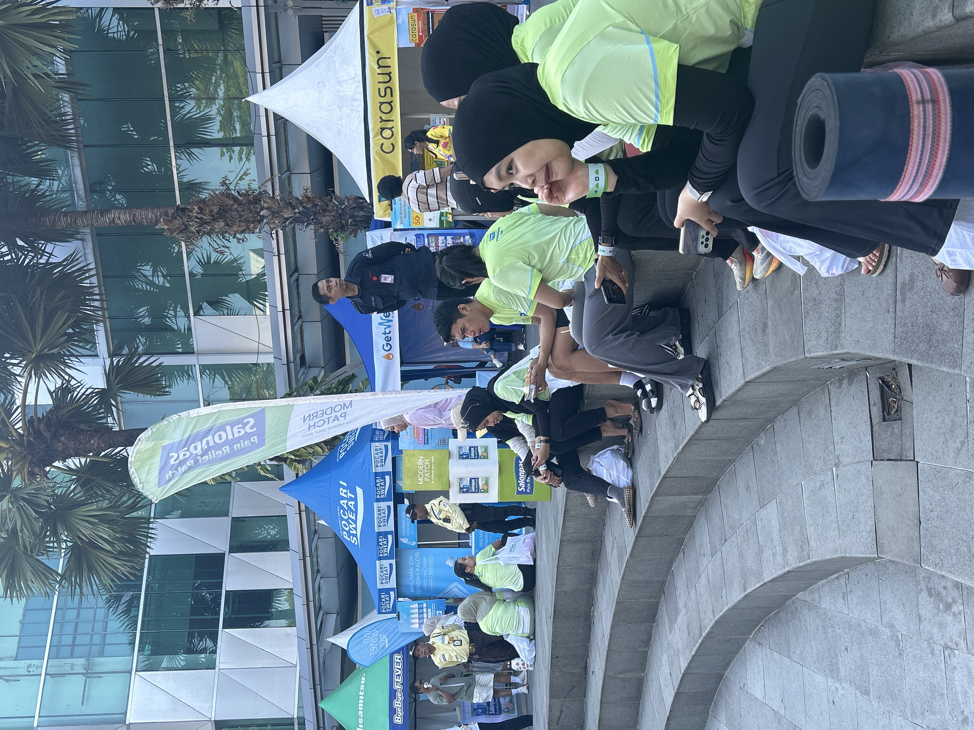 In Makassar, Indonesia, at an outdoor event, several people are seated on curved stone steps. Many are wearing matching light green t-shirts with blue and yellow accents. To the left, some individuals are standing, including two men in yellow security uniforms and another in a black security uniform. Several tents are visible in the background, including blue tents with "POCARI SWEAT" branding and a yellow tent with "carasun" branding. Palm trees and a modern building with glass windows are also present. Some people are holding smartphones, and a rolled-up yoga mat is visible in the foreground. The scene appears to be a public gathering or promotional event.