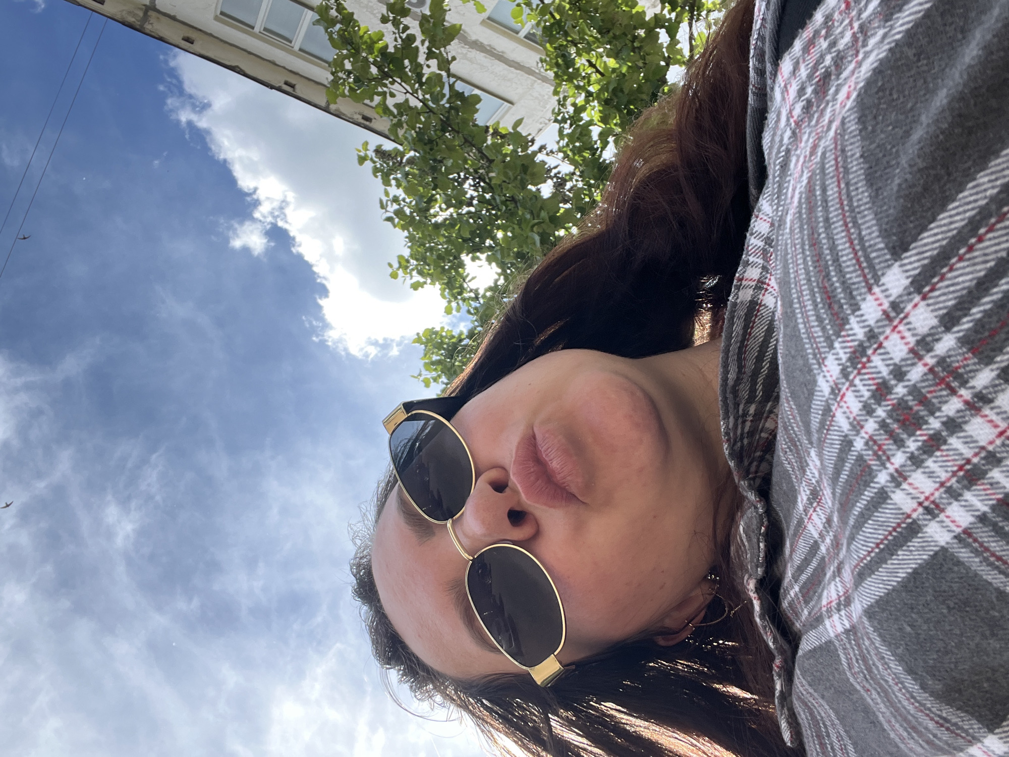 An outdoor, low-angle selfie captures a person looking upwards against a bright blue sky in Noviy, Russia. The individual, seen from the chest up, has long, dark hair and wears gold-framed sunglasses with dark lenses. Their lips are slightly pursed, forming a casual or playful expression. They are dressed in a grey flannel shirt featuring white and red plaid patterns, worn over a dark top. A small earring is visible on their left ear.
The background reveals a sunny daytime scene with scattered white clouds across the sky. In the upper right section of the image, the facade of a light-colored building with windows is partially visible, somewhat obscured by green tree leaves and branches. A thin wire or power line is noticeable against the sky in the upper left, and a small, dark, bird-like object is also seen in the distance. The overall impression is one of a bright, casual moment outdoors. There is no visible text.
