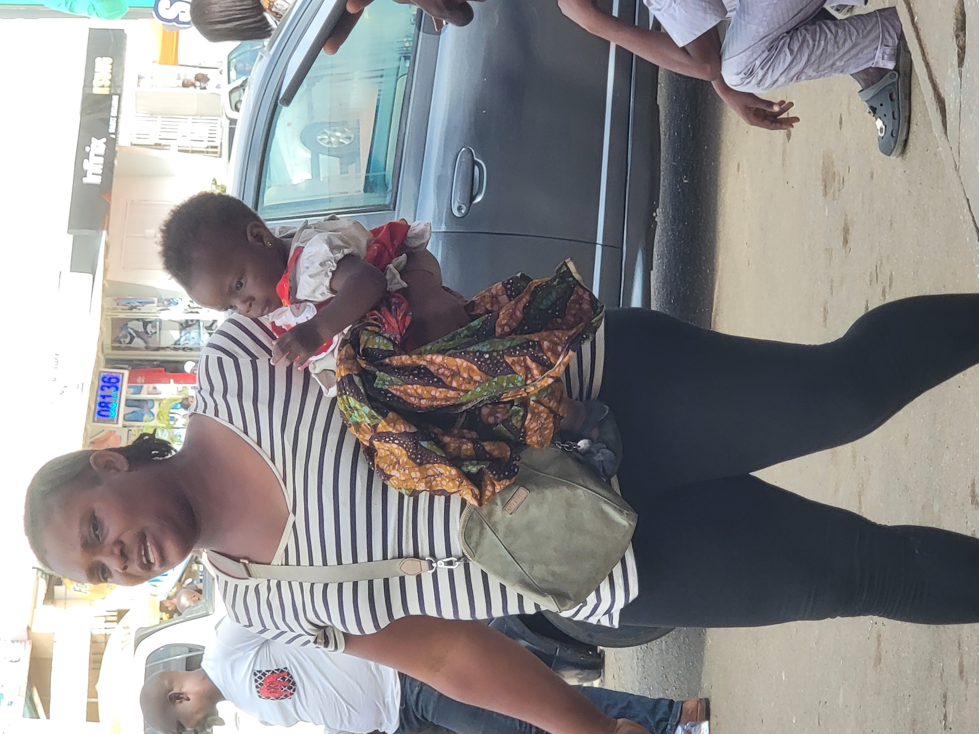 The image captures an outdoor street scene in daylight, featuring a dark-skinned woman carrying a baby on her back. The woman, positioned centrally, is wearing a black and white striped short-sleeved top, black leggings, and a light beige crossbody bag slung across her chest. She has a subtle smile and dark hair pulled back. The baby, with dark skin and dressed in a white top with red patterns, is wrapped in a vibrant, multi-colored fabric and appears to be looking downwards over the woman's shoulder.
In the background to the left, a white building displays a prominent black sign with the text "Infinix". Below this, a blue digital-like display shows "08136". To the right, a dark grey car is partially visible, with a person wearing light-colored clothing and dark Crocs-style shoes standing nearby, their arm and lower leg visible. Another man, wearing a white t-shirt with a patterned emblem and blue jeans, is partially visible behind the central woman. The ground is paved, suggesting a sidewalk or urban pathway. The bright lighting indicates it is daytime and sunny. The image is oriented sideways, with the subjects appearing rotated 90 degrees from an upright position.