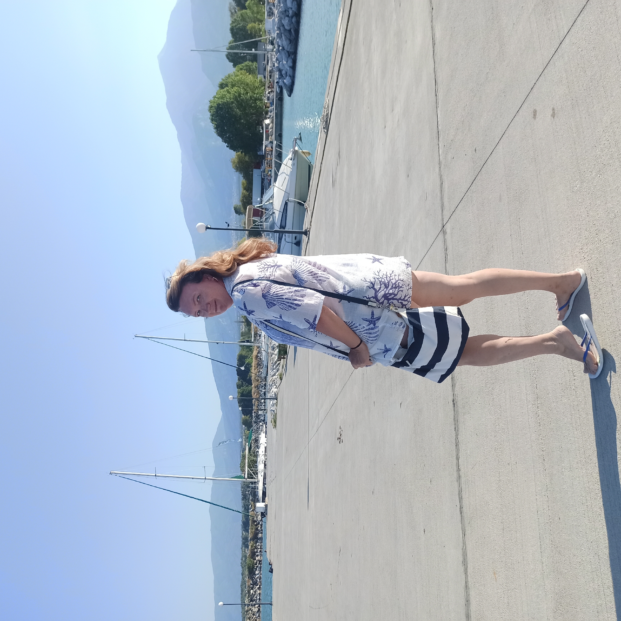 A woman stands on a concrete pier in Limani Litochorou, Greece, facing away from the camera and looking over her shoulder. She is wearing a white shirt with a blue seashell print, a striped tote bag slung over her shoulder, and flip-flops. The pier is bordered by a calm blue body of water on the right, where several white boats and sailboats are docked. Rocky jetties and trees line the water's edge, with a range of hazy blue mountains visible in the background under a clear, bright sky. The scene is bathed in the warm light of what appears to be late afternoon.