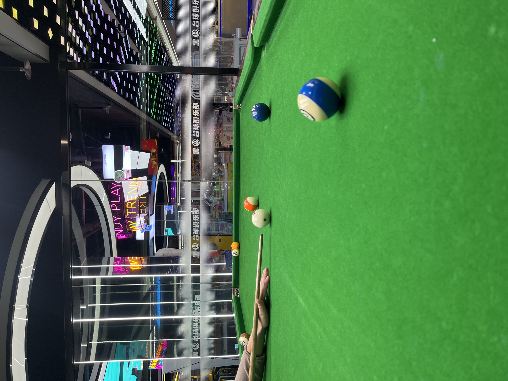 This media file captures a close-up, low-angle view of a billiard table with a green felt surface. Several billiard balls are scattered across the table, including a blue ball, a blue and white striped ball, an orange ball, and a white ball. A person's hand, holding a cue stick, is visible in the foreground, poised to strike one of the balls.
The background is a blur of activity, suggesting an indoor entertainment venue. Neon lights and electronic displays with Chinese text are visible, indicating a commercial or amusement area. The text includes "ENDY PLAY," "AYTREND," and "YUJANCHAO," along with repeated instances of "黑台球俱乐部" (hēi táiqiú jùlèbù), which translates to "Black Billiards Club." The lighting suggests it could be evening or a dimly lit indoor space. The scene conveys a sense of leisure and recreation, with the focus on the game of billiards. The presence of neon signs and the mention of a "billiards club" place the setting in a vibrant entertainment district.