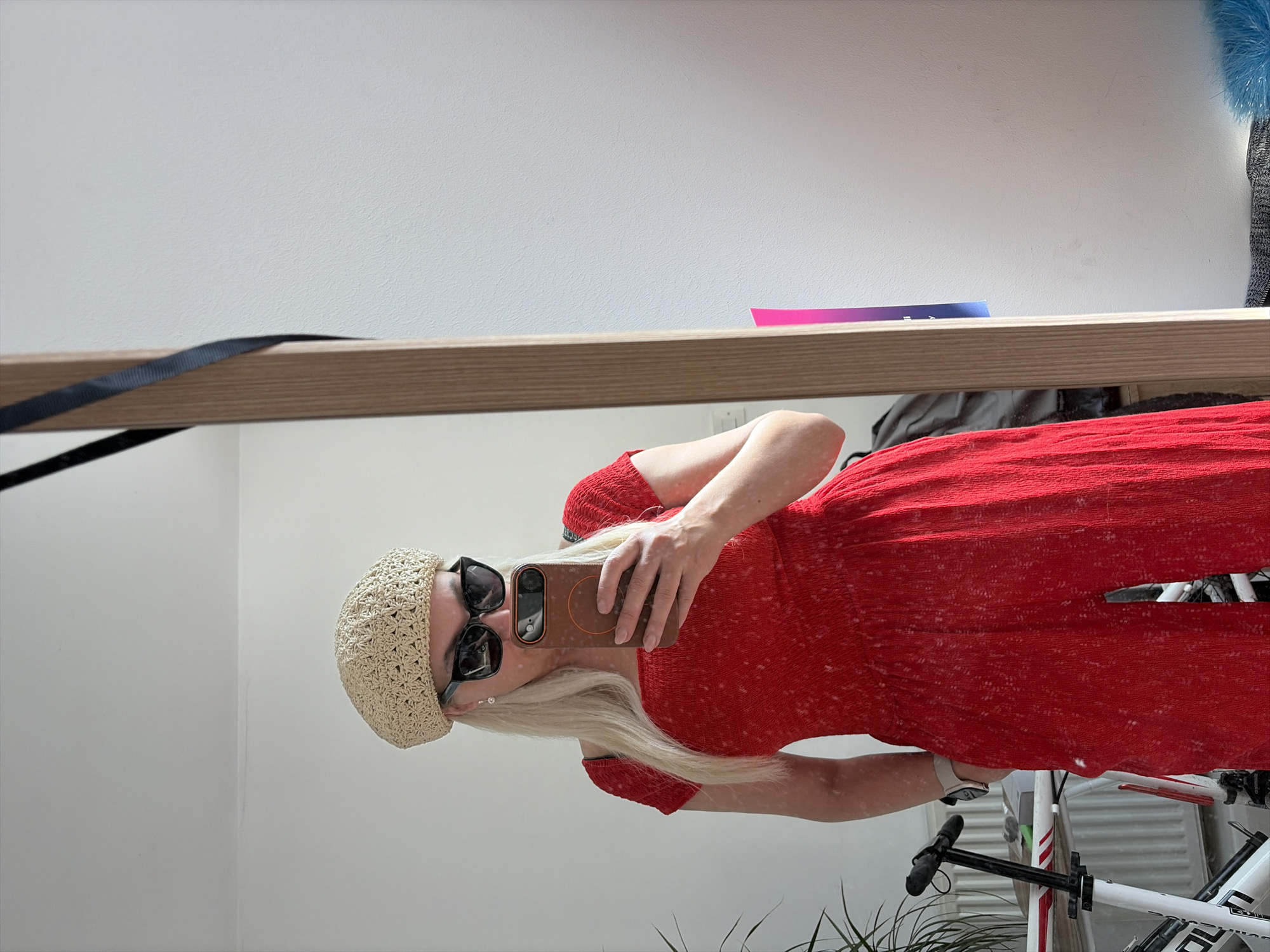 The image is a selfie taken in a mirror. The subject is a person wearing a red jumpsuit, a beige crocheted beret, and sunglasses. They are holding a phone in their hand. The background shows a white wall and a portion of a bicycle. The location cannot be determined as Salford, United Kingdom based on the visual information.