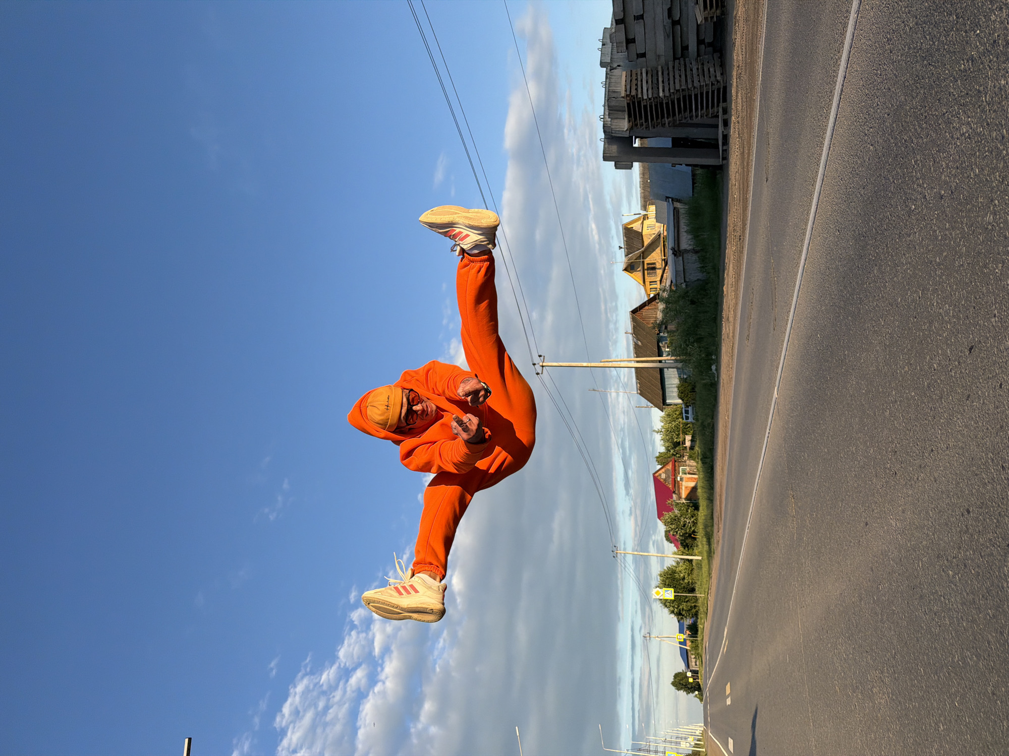 A person is captured mid-air, performing a dynamic jump or kick against a bright blue sky with scattered white clouds. The subject, an individual with a beard, wears a vibrant orange tracksuit consisting of a hoodie and sweatpants, an orange beanie, and light-colored sneakers with visible red stripes, indicative of the Adidas brand. One leg is extended high in a kick, while the other is bent, and both arms are flexed with hands clenched into fists. Sunglasses obscure part of the person's face.
The scene is set outdoors in a semi-urban or rural area of Chaikovsky, Russia, during what appears to be late afternoon or early evening, characterized by soft, warm sunlight. A paved road with a distinct white dividing line occupies the foreground. Along the sides of the road, various houses with traditional architecture and some green trees and bushes are visible. On the left side of the road, large stacks of concrete slabs or construction materials are piled high. Power lines crisscross the upper sky. Several yellow diamond-shaped road signs are present, including one depicting a pedestrian crossing symbol and another with an exclamation mark, indicating a warning. The image conveys a sense of energetic movement within an everyday landscape.