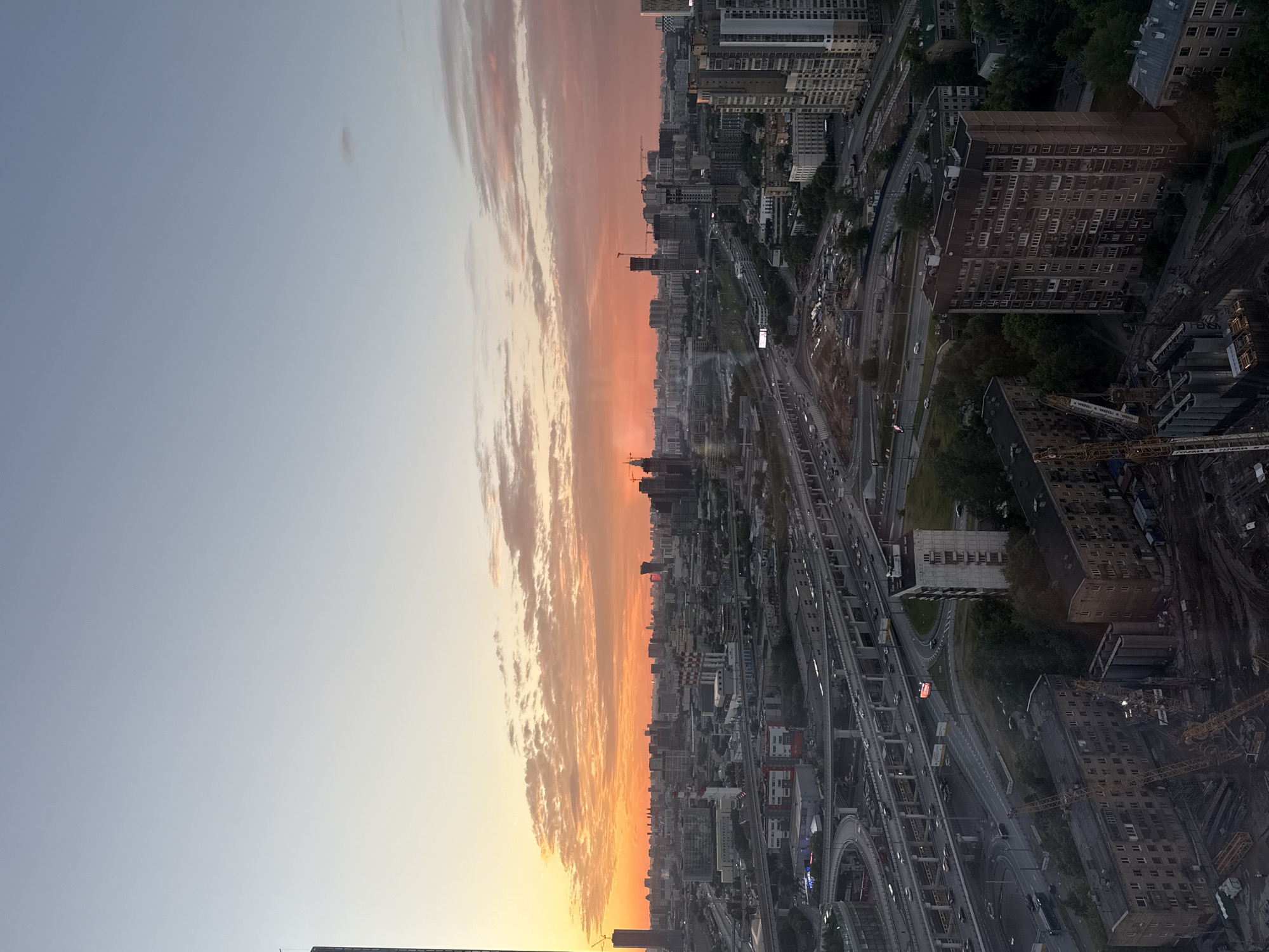 This is an aerial, wide-angle photograph of a city skyline at dusk, likely taken from a high vantage point. The sky is dominated by a vibrant sunset, with hues of orange, pink, and yellow illuminating the clouds and casting a warm glow over the cityscape. The lower half of the image is filled with a dense urban environment characterized by numerous buildings of varying heights and architectural styles, including residential apartment blocks, high-rise office buildings, and industrial structures with chimneys. A complex network of multi-level highways and overpasses with visible car traffic cuts through the urban landscape, indicating a significant transportation hub. Construction cranes are also visible, suggesting ongoing development in some areas of the city. The photograph captures a sprawling metropolis, with the details becoming less distinct in the distance. The overall impression is one of a busy, modern city bathed in the dramatic light of sunset. Based on the visual cues, such as the architectural styles of the buildings and the urban infrastructure, the location is identified as Khimki, Russia.
