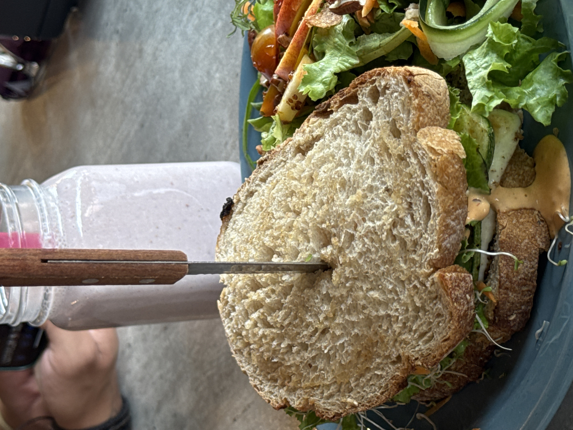 A high-angle, close-up shot depicts a partially eaten sandwich pierced by a knife, resting on a blue plate alongside a salad. A glass jar, likely containing a smoothie or beverage, lies near the sandwich. The location is suggested to be in Tlalixtac de Cabrera, Mexico. The sandwich consists of whole-grain bread, sprouts, and some type of sauce. The salad contains lettuce, carrots, and apple slices. The knife is partially inserted into the bread, obscuring its fillings. The photo seems to have been taken indoors, possibly in a restaurant or cafe. An unidentifiable person's hand is at the bottom left corner of the photo.