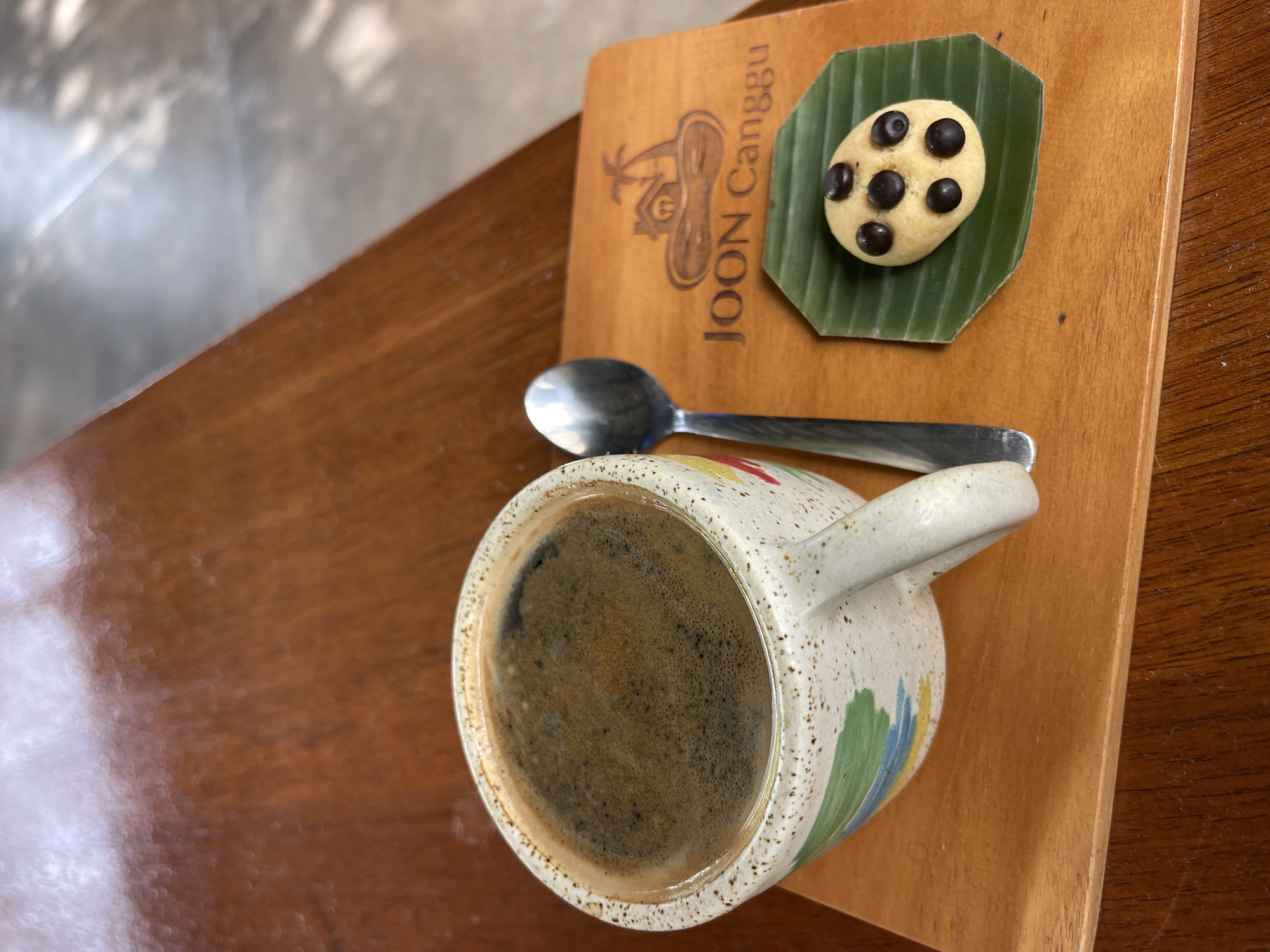A close-up, overhead shot captures a coffee cup filled with dark coffee, its rim speckled with dark specks. Beside it, a metal spoon rests on a wooden board that features a logo of a house with palm trees and the text "JOON Canggu". A single chocolate chip cookie sits on a green banana leaf atop the wooden board. The coffee and cookie are situated on a dark wooden table. The scene appears to be in a cafe in North Kuta, Indonesia, indicated by the "JOON Canggu" branding.