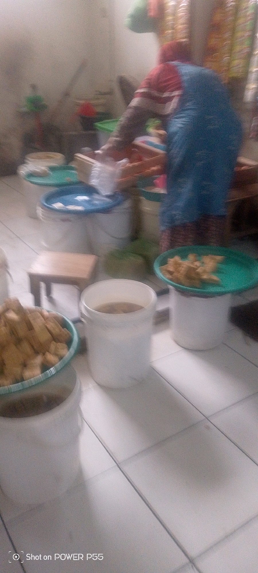A blurry, medium shot captures an indoor scene with a person wearing a red and blue outfit. The person is standing near a wooden crate filled with items, possibly food. Around them are several large white buckets, some with blue lids, and green plates holding food that appears to be fried. A small wooden stool is visible to the left. The floor is tiled. The background shows a wall with curtains and shelves with objects, suggesting a kitchen or preparation area. The overall lighting is dim.