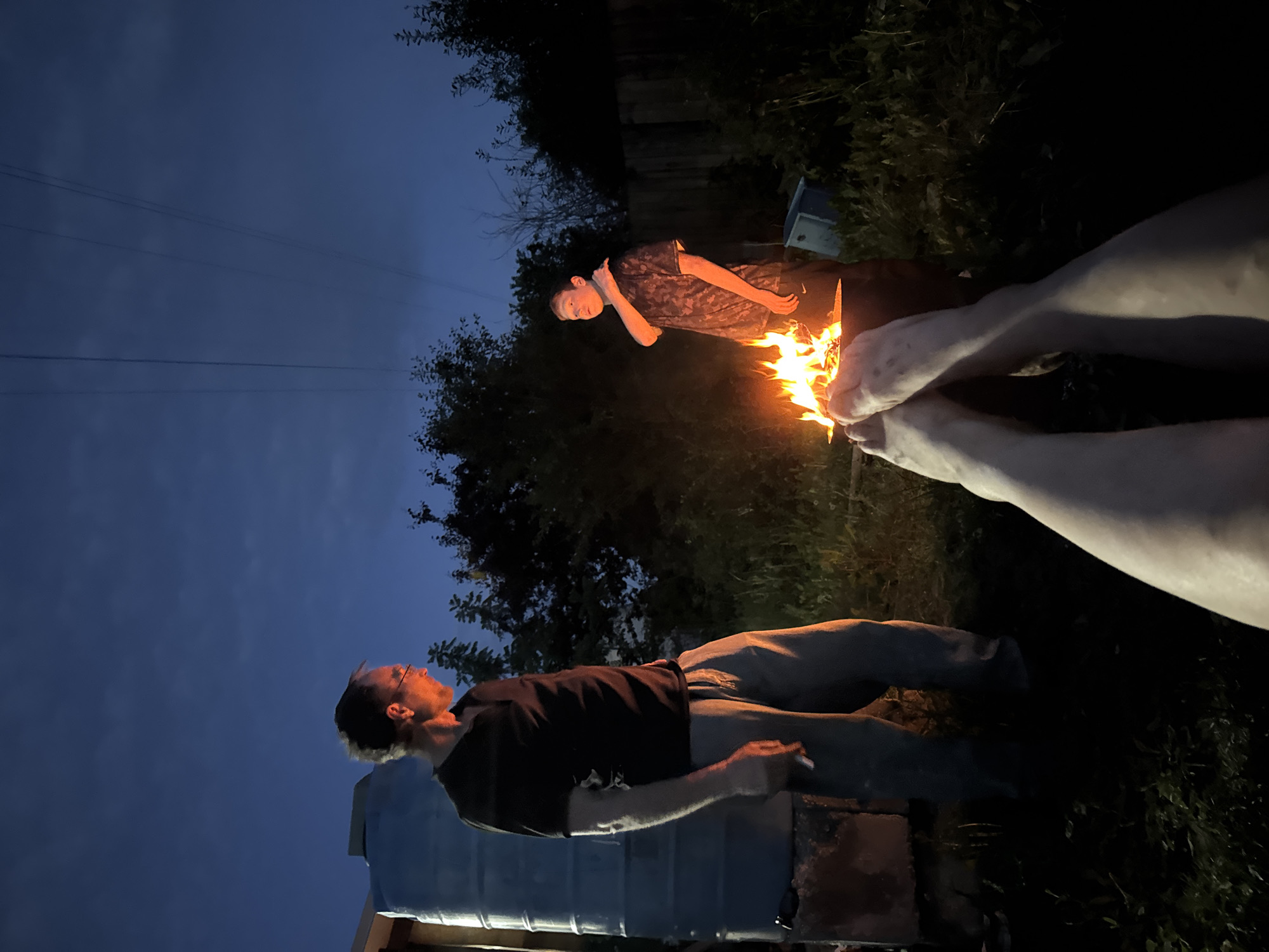 In the twilight, two individuals stand near a bonfire. On the left, a man with glasses, wearing a dark t-shirt and light-colored pants, holds a cigarette in his right hand. Beside him is a large, cylindrical blue water tank. On the right, a young person in a patterned t-shirt and dark pants looks towards the fire. The setting appears to be a backyard, with overgrown grass, trees, and a wooden fence visible in the background. Overhead, power lines stretch across the dark blue sky. The foreground shows a pair of bare legs, suggesting the perspective of someone sitting or lying down, looking at the scene. The image captures a moment of outdoor gathering, possibly in Chaikovsky, Russia, given the context.