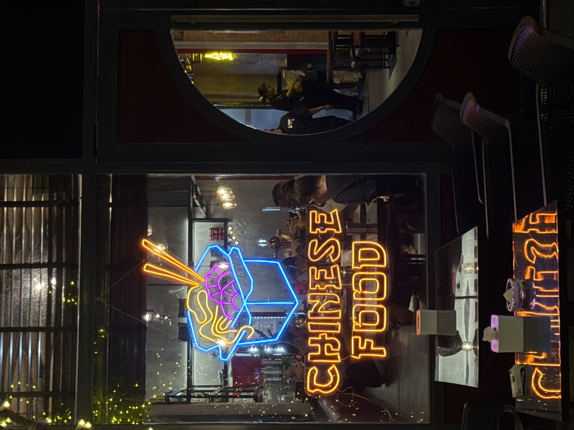 A night scene captured in Đà Nẵng, Vietnam, shows the reflection of a "CHINESE FOOD" neon sign and a noodle box sign in the window of a Chinese restaurant. The restaurant appears dimly lit, with a glimpse of patrons dining inside, visible through the window reflection. Chairs are visible, as well as tables and decorative lights. A round window with people in it is visible above the restaurant window.