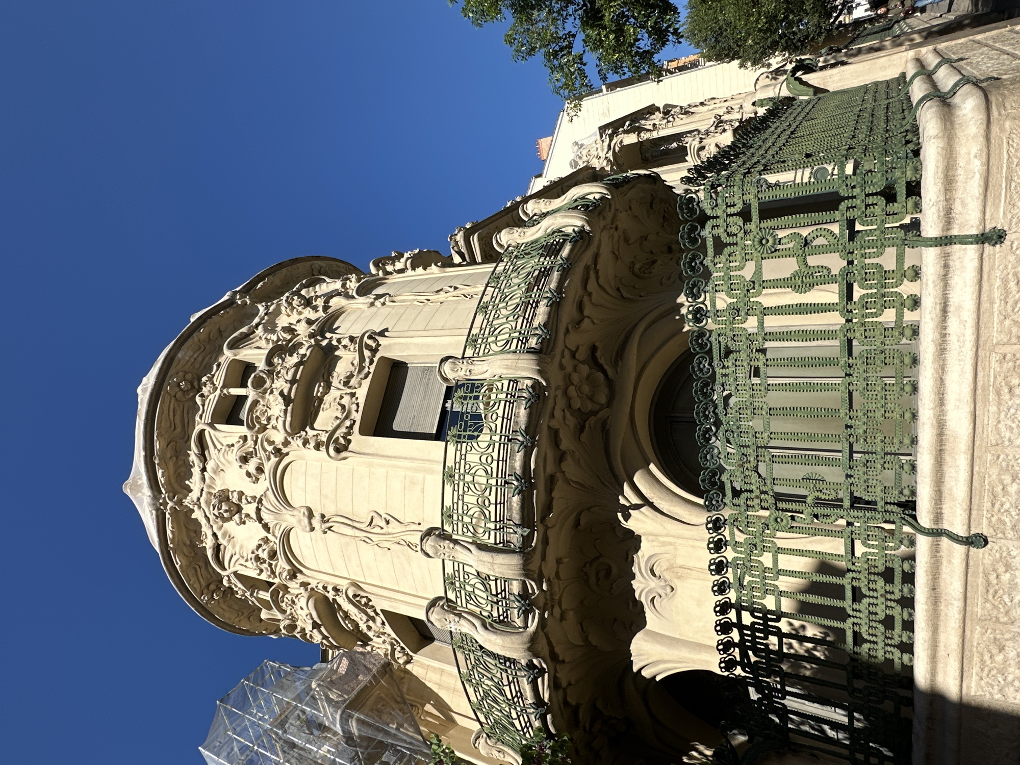 The image is a low-angle shot of a highly ornate building in Madrid, Spain. The building has a turret-like structure on the corner, with elaborate carvings and sculptural details on the facade. The stonework is light beige. There are windows of different shapes and sizes, some with green metal latticework or grilles. Two balconies are visible, with intricately designed green iron railings. Scaffolding covered with a net is visible on the lower left of the frame. A clear blue sky fills the upper left corner of the picture, while some greenery from trees appears at the top center. The building's architecture appears to be in the Art Nouveau or Modernista style.