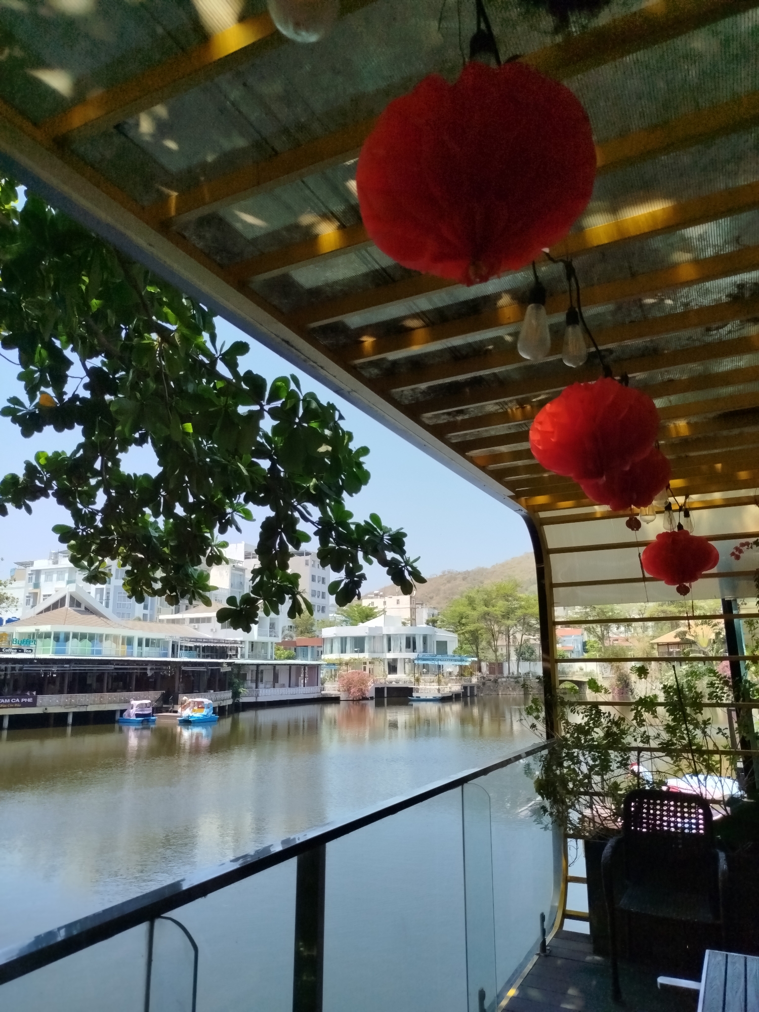 The image depicts an outdoor scene overlooking a body of water, likely a lake or river, with buildings visible in the background. In the foreground, there is a seating area with chairs and plants, suggesting a restaurant or cafe. Red paper lanterns hang from the ceiling, providing a decorative element. The view extends to a waterfront area with buildings and trees. In the background, there are white buildings, possibly residential or commercial structures. There are no people visible in the image. Based on the architecture and the presence of red lanterns, it is possible that this scene is located in a tourist area within Ho Chi Minh City, Vietnam, though specific landmarks would be required for verification.